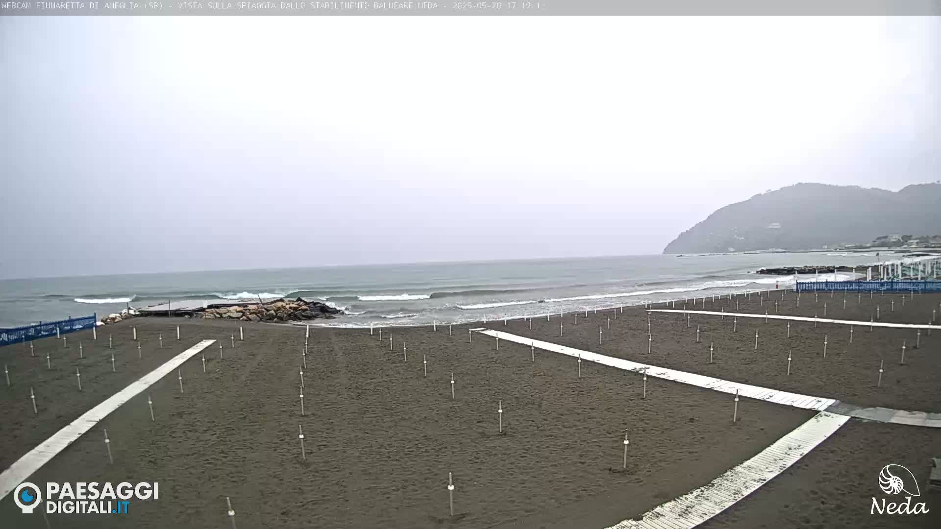 A dark-sand beach with rows of posts and walkways is shown under an overcast sky, with a calm sea and a distant mountain visible in the background.