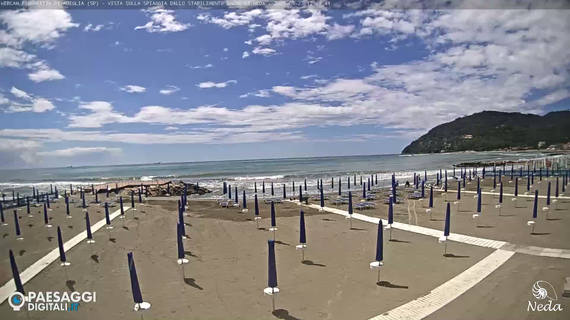 A sandy beach with numerous closed blue umbrellas under a partly cloudy sky and a calm sea with small waves in the background.