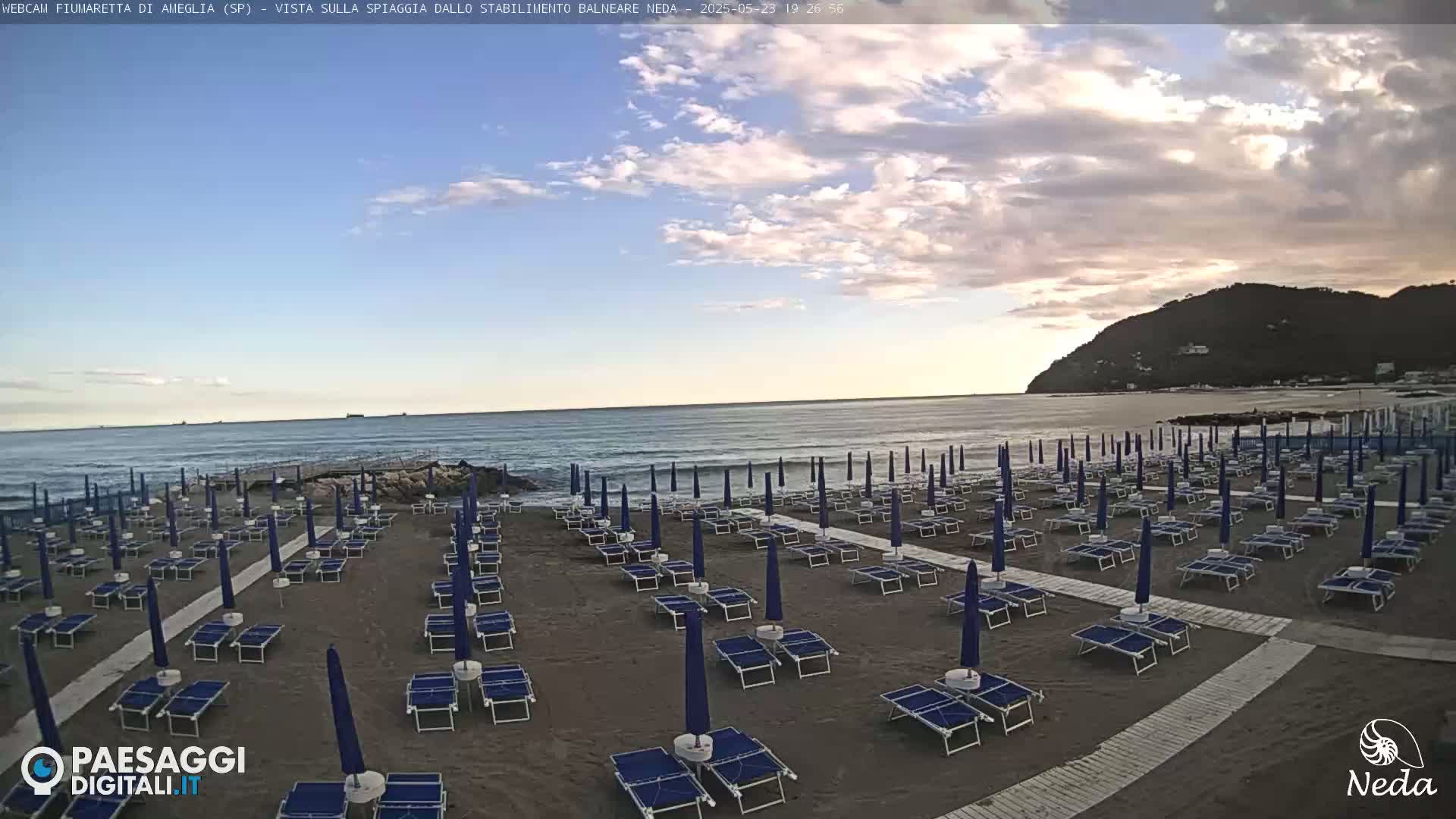 A beach with rows of closed umbrellas and lounge chairs sits under a partly cloudy sky next to a calm ocean and a distant hill.