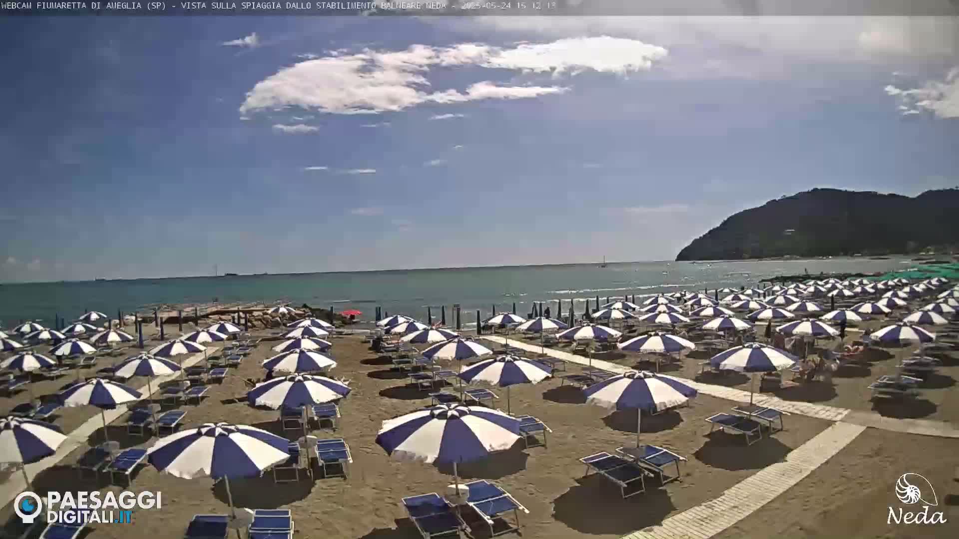 A sandy beach with numerous blue and white striped umbrellas and lounge chairs is visible under a partly cloudy sky, overlooking a calm sea and a distant mountain.