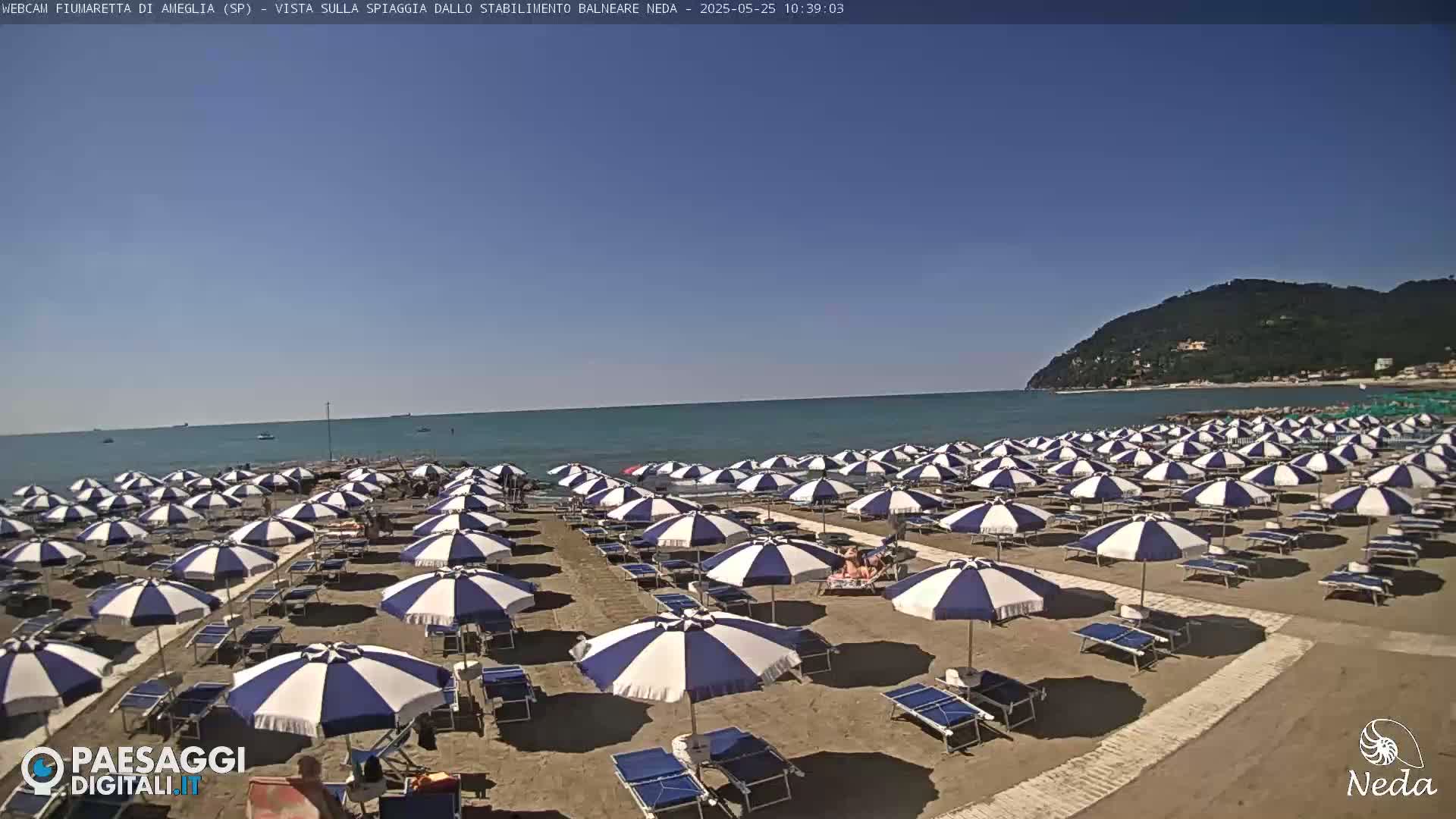 A sandy beach with many blue and white striped umbrellas under a clear blue sky and calm ocean.