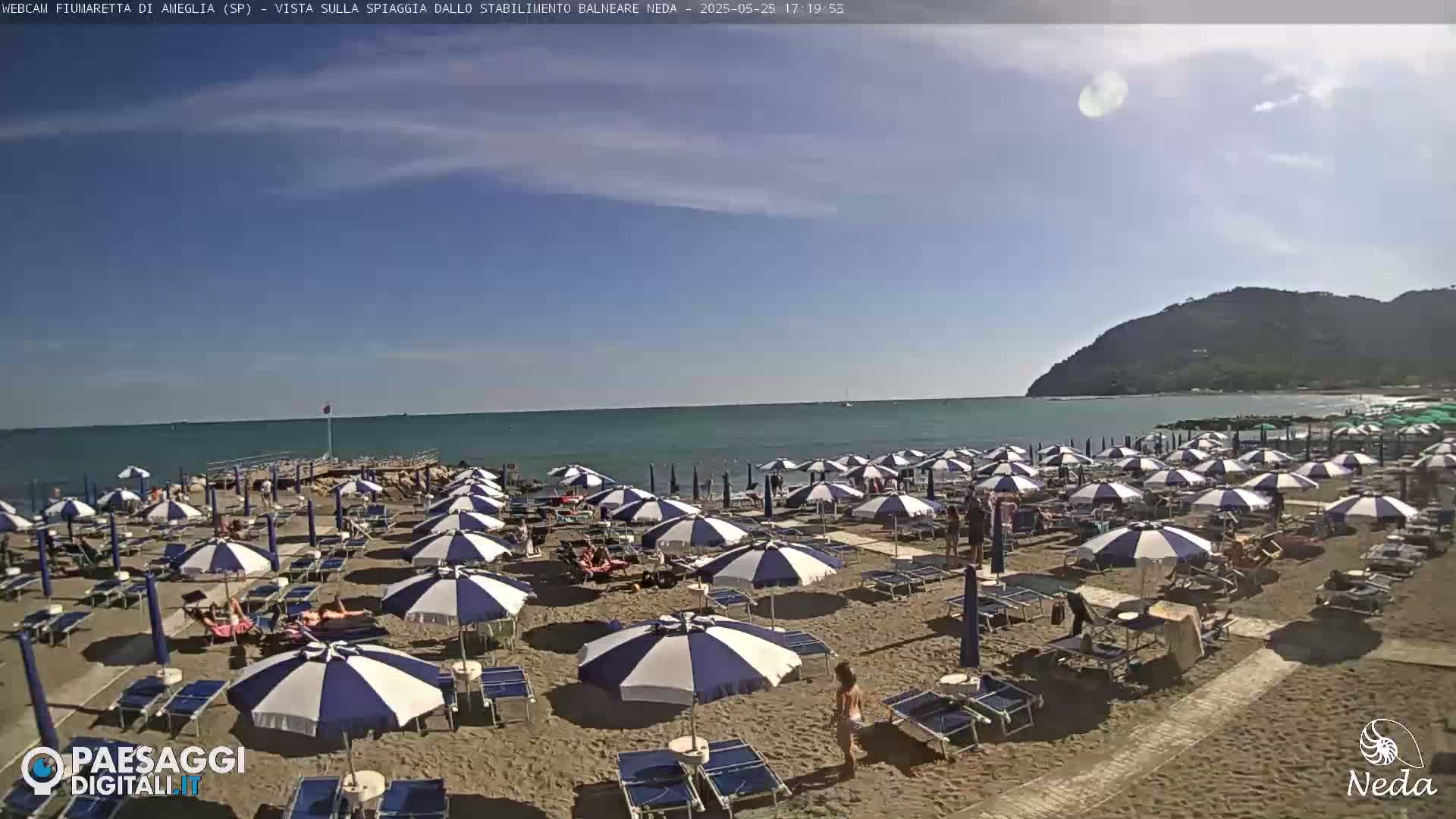 A sandy beach with numerous blue and white striped umbrellas and beach chairs is situated next to a calm ocean under a mostly clear sky, with a hill visible in the background.