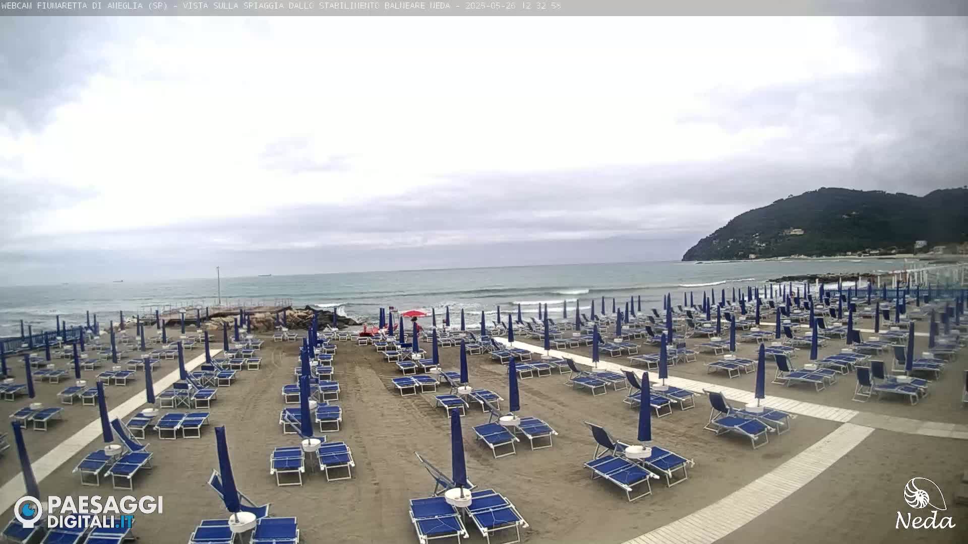 A mostly cloudy day at a beach with rows of closed blue umbrellas and lounge chairs on sandy shore, and a calm ocean with small waves in the background.