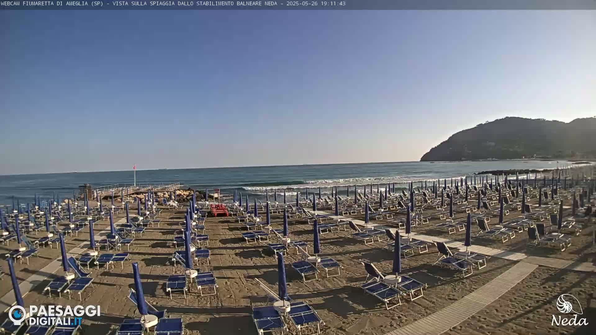 A sandy beach with rows of closed beach umbrellas and lounge chairs sits beside a calm ocean under a clear blue sky, backed by a distant hill.