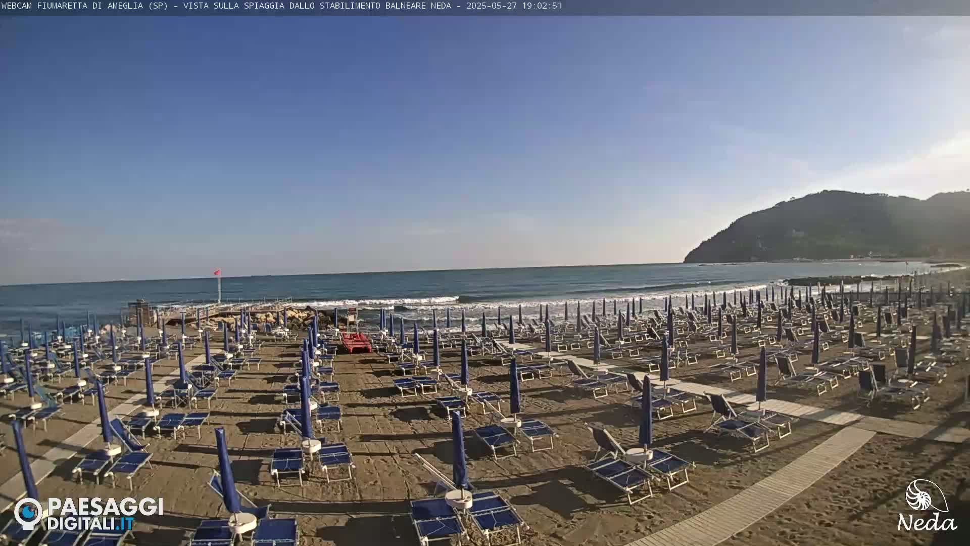 A sandy beach with rows of closed beach umbrellas and lounge chairs sits on a calm, sunny ocean under a clear blue sky, with a distant hill visible on the right.