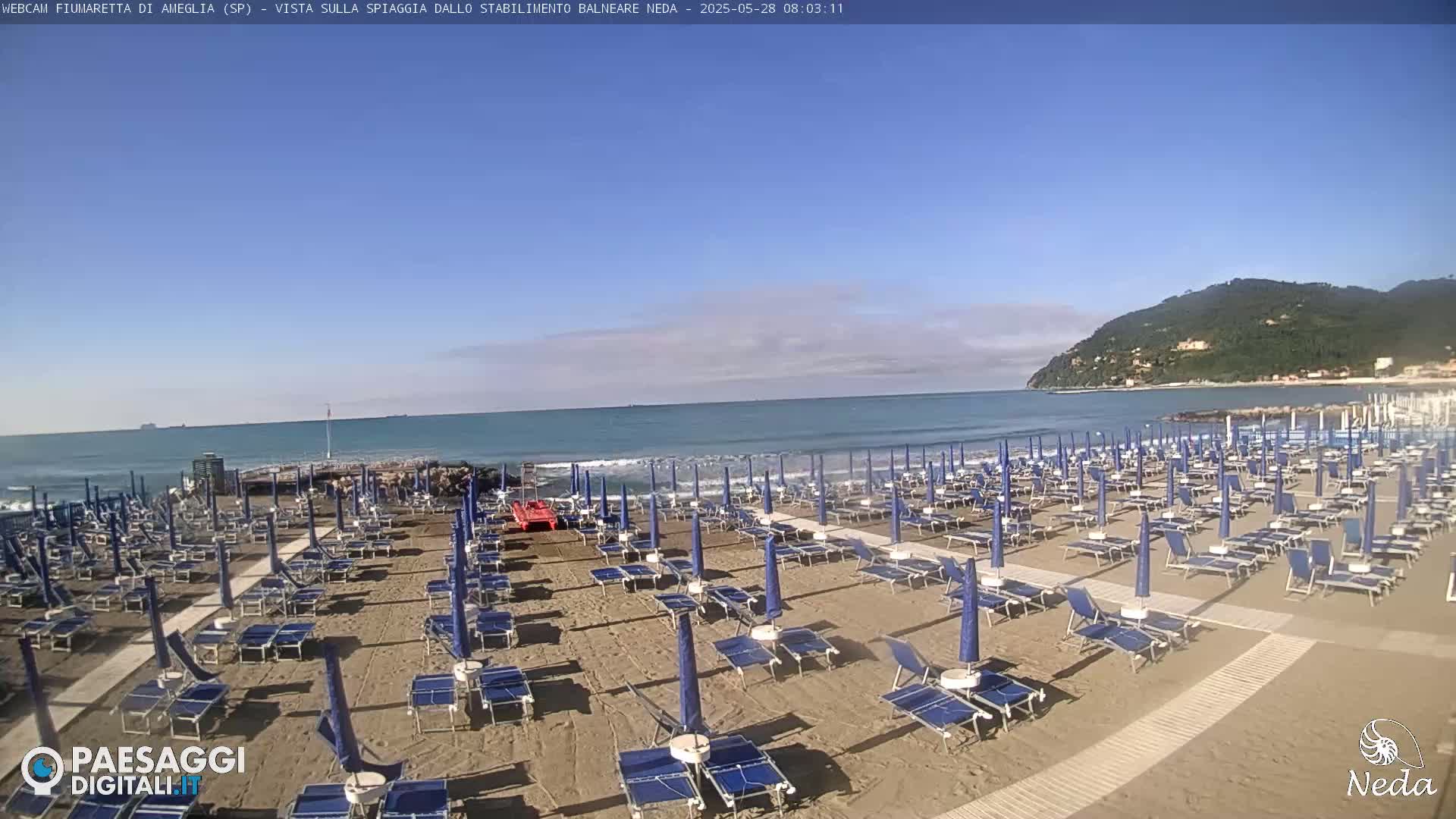 A sandy beach with numerous blue beach umbrellas and lounge chairs is situated next to a calm ocean under a mostly clear sky, with a green hill visible in the distance.