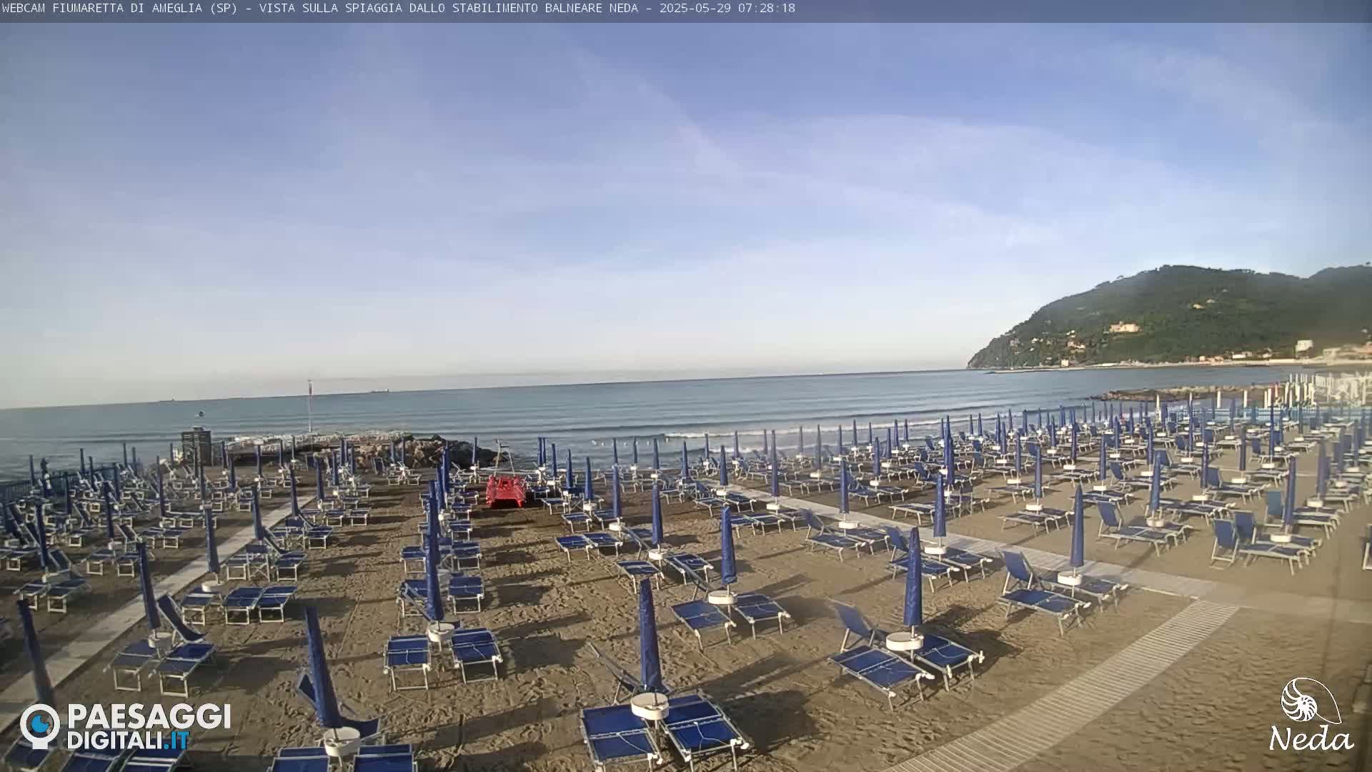 A sandy beach with rows of blue beach umbrellas and lounge chairs is visible, under a mostly clear sky, next to a calm ocean with a mountain in the background.