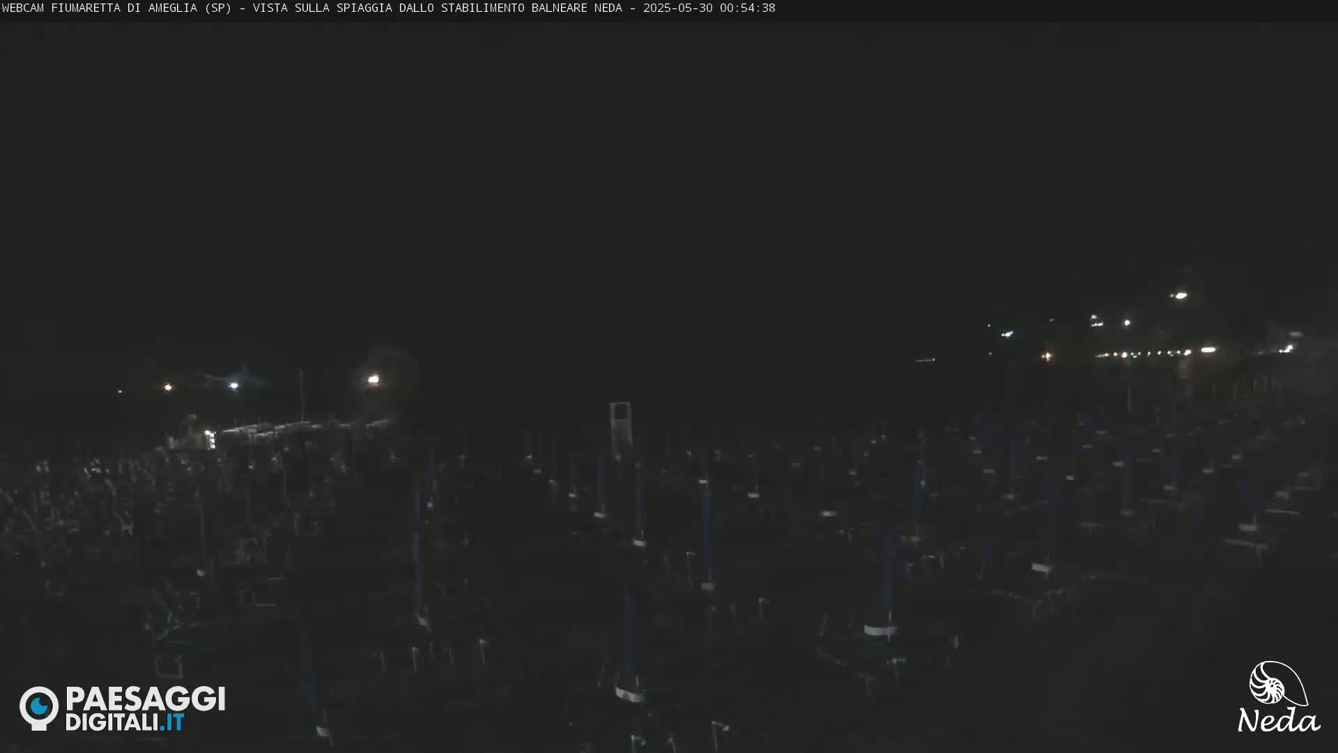 At night, a beach is mostly dark except for numerous dark objects, possibly beach chairs, sparsely scattered across the sand, with some lights visible in the distance.