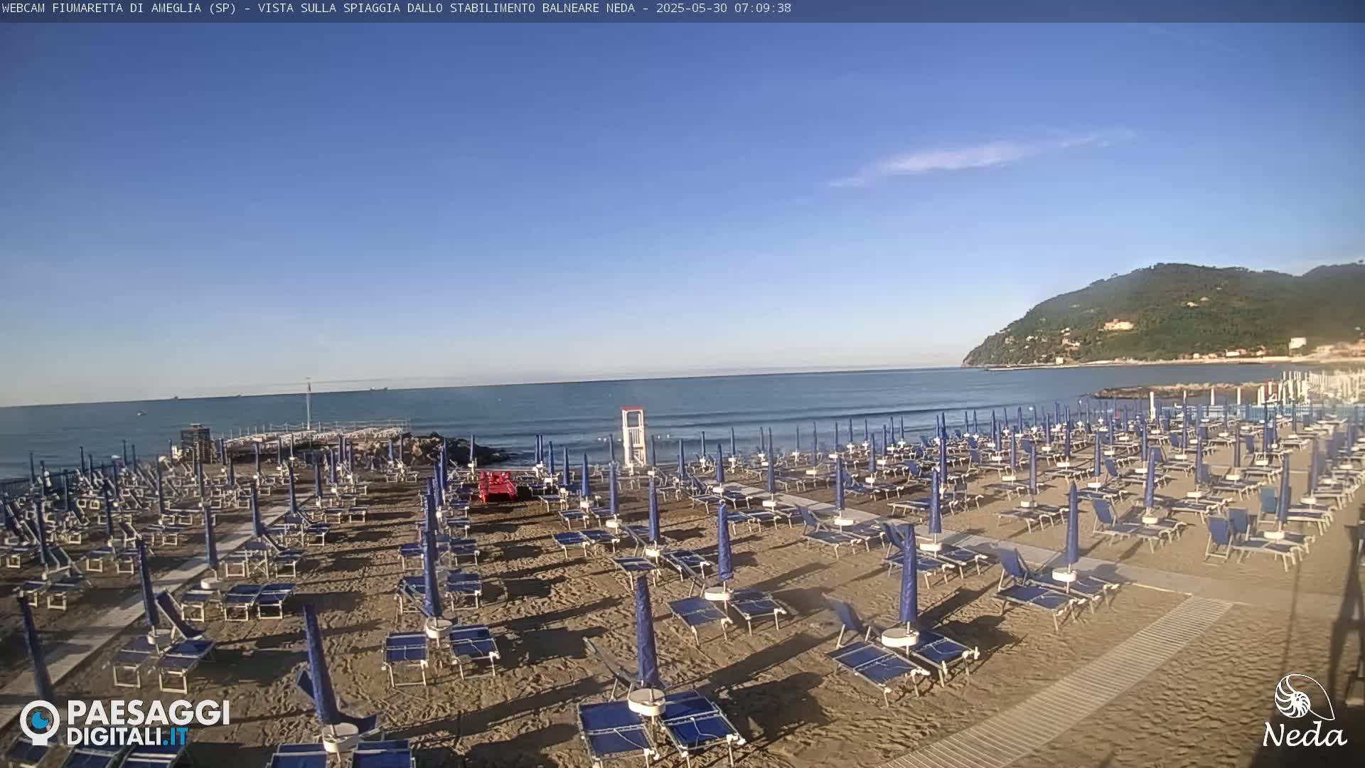 A sandy beach with rows of closed blue umbrellas and lounge chairs sits beside a calm, blue ocean under a clear sky, with a green hill visible in the distance.