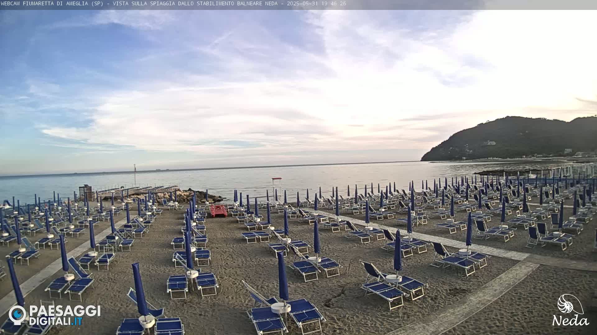 A sandy beach with numerous blue beach umbrellas and lounge chairs is visible, under a partly cloudy sky, overlooking a calm sea and a distant hill.