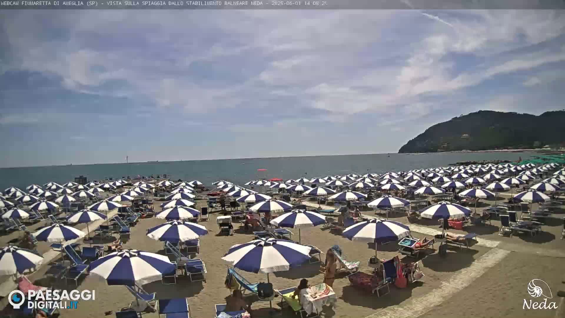A sandy beach is crowded with many blue and white striped umbrellas under a partly cloudy sky.