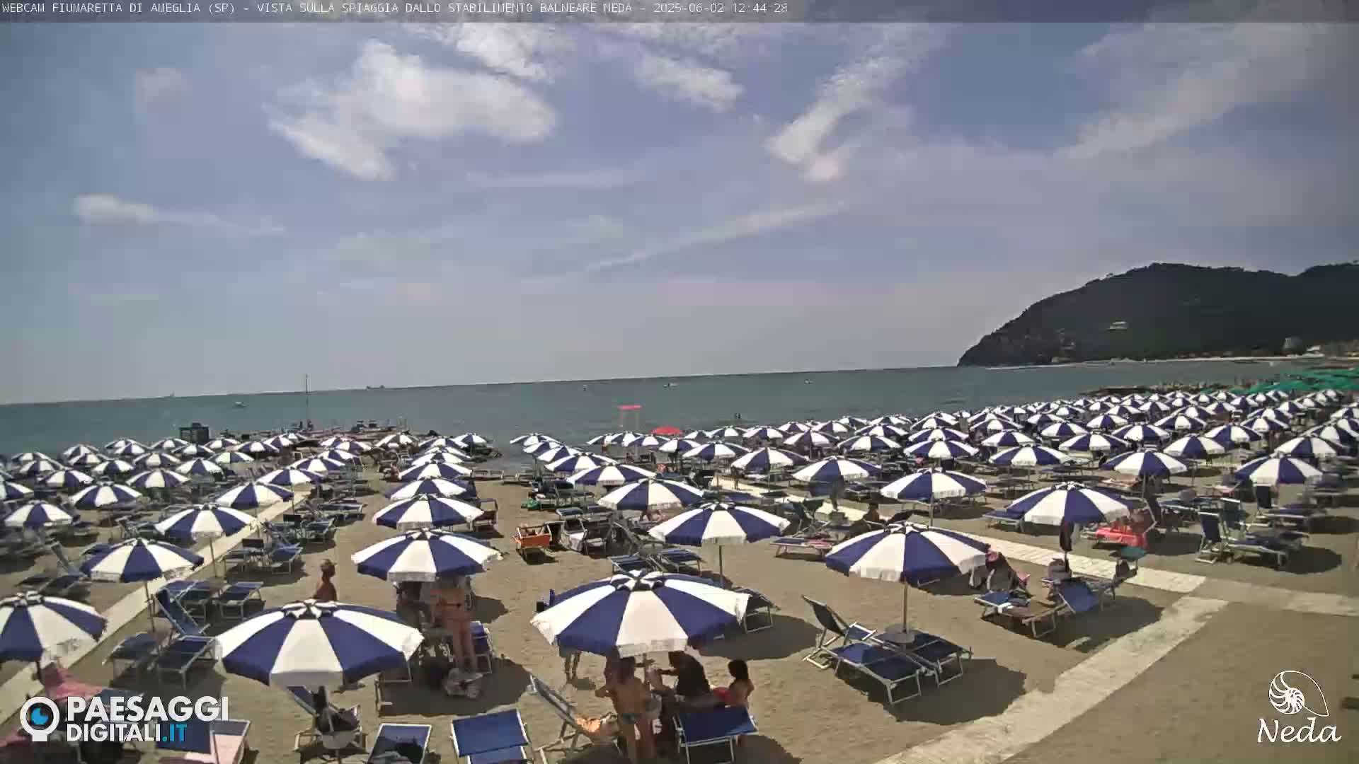 A sandy beach filled with numerous blue and white striped beach umbrellas under a partly cloudy sky, with a calm sea in the background and a hill visible in the distance.