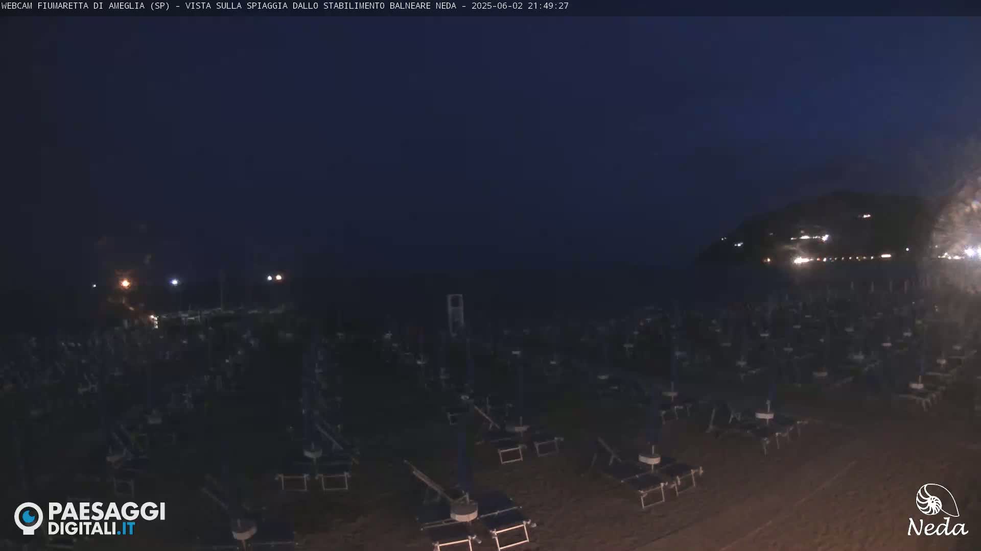 A dark beach at night is filled with numerous closed beach umbrellas and lounge chairs, with a dimly lit hillside visible in the distance.