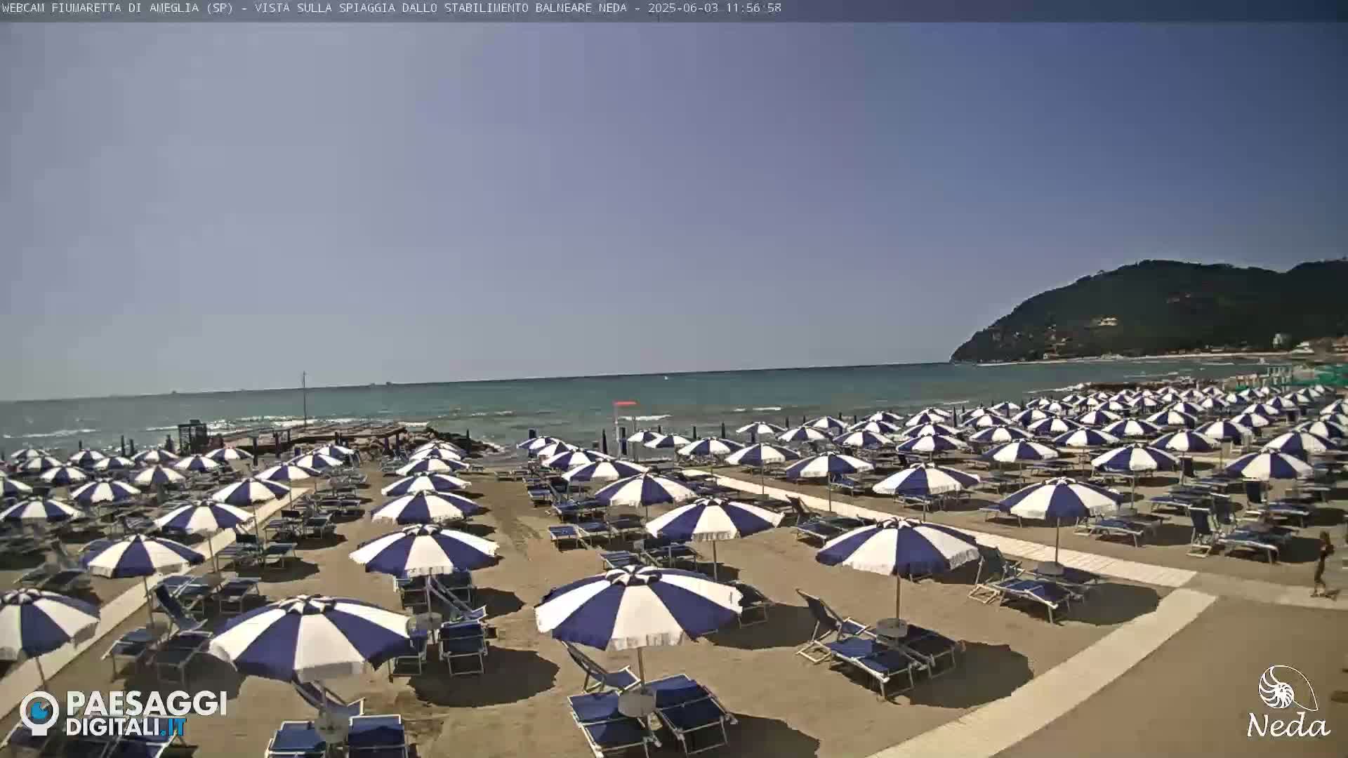 A sandy beach with numerous blue and white striped umbrellas and lounge chairs stretches to a calm sea under a clear, sunny sky, with a distant hillside visible.