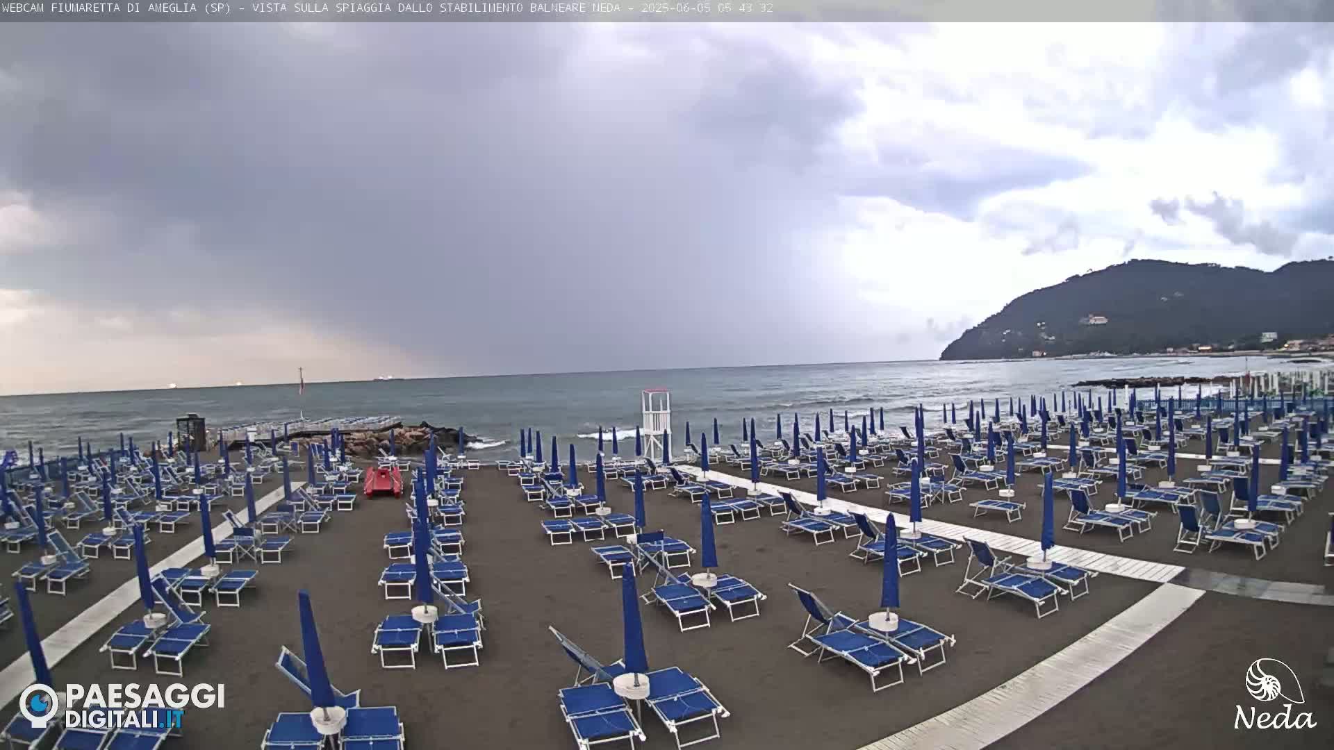 A mostly cloudy day shows a beach with rows of closed blue umbrellas and lounge chairs on dark sand, a lifeguard stand, and a calm sea with small waves in the background.