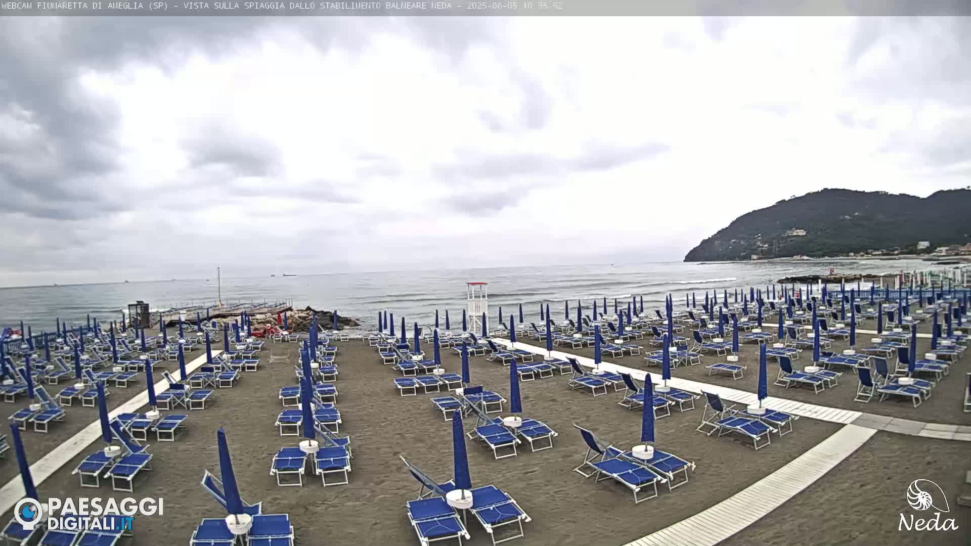A mostly cloudy day at a beach with rows of blue umbrellas and lounge chairs on a sandy shore, and a calm ocean.