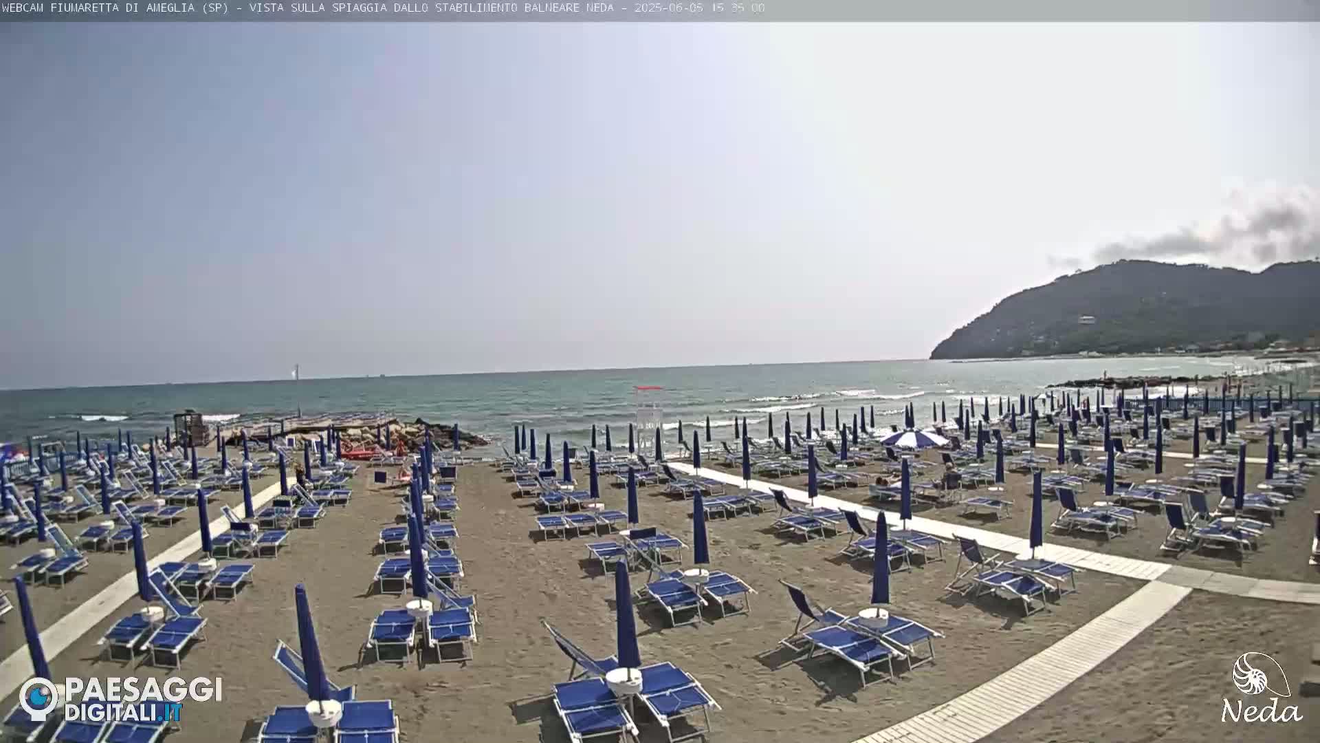A mostly sunny beach with numerous rows of blue beach umbrellas and lounge chairs situated next to a calm ocean and a distant mountain.
