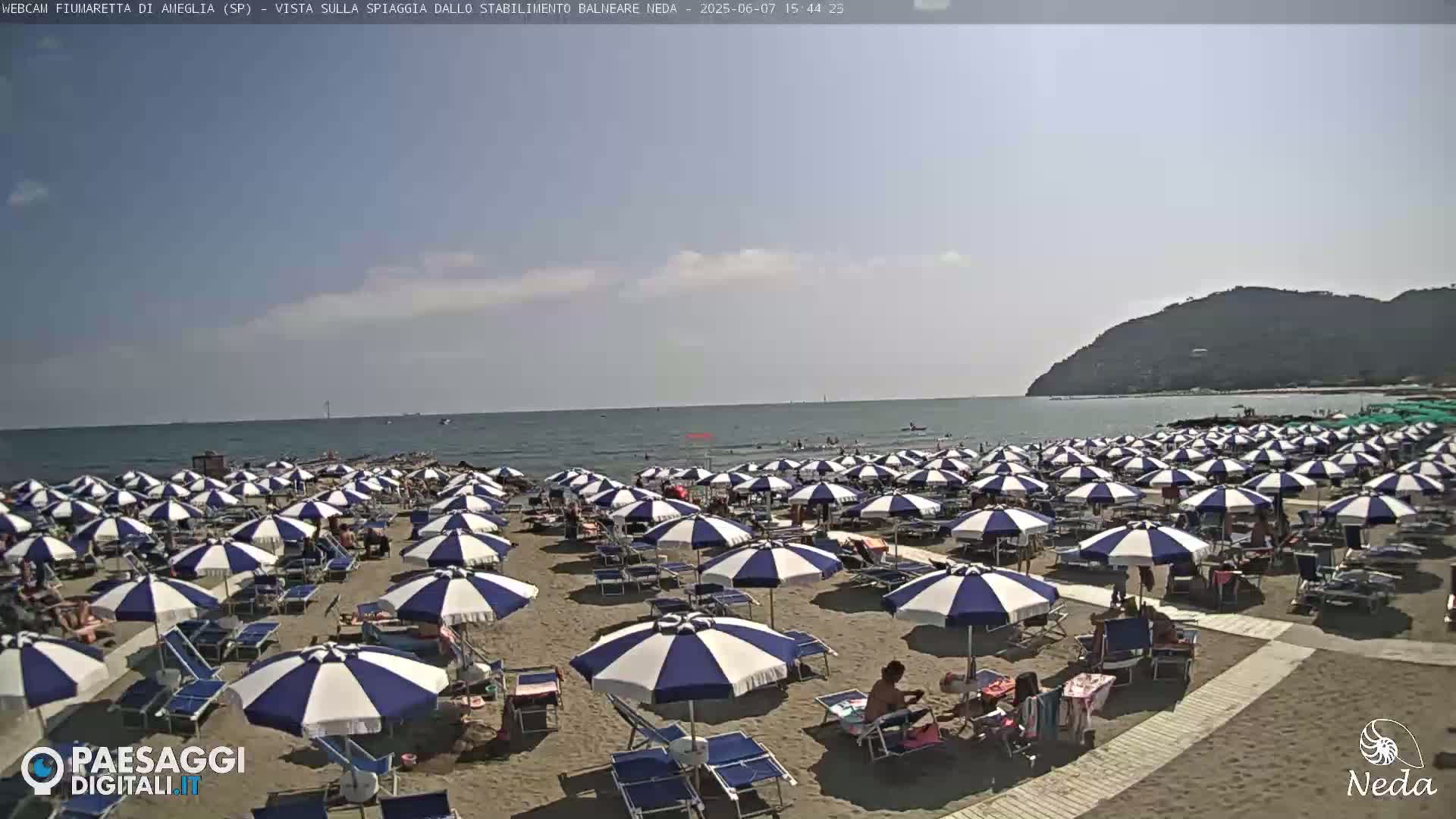 A sandy beach with many blue and white striped umbrellas and lounge chairs under a mostly clear sky, next to a calm ocean and a distant hill.