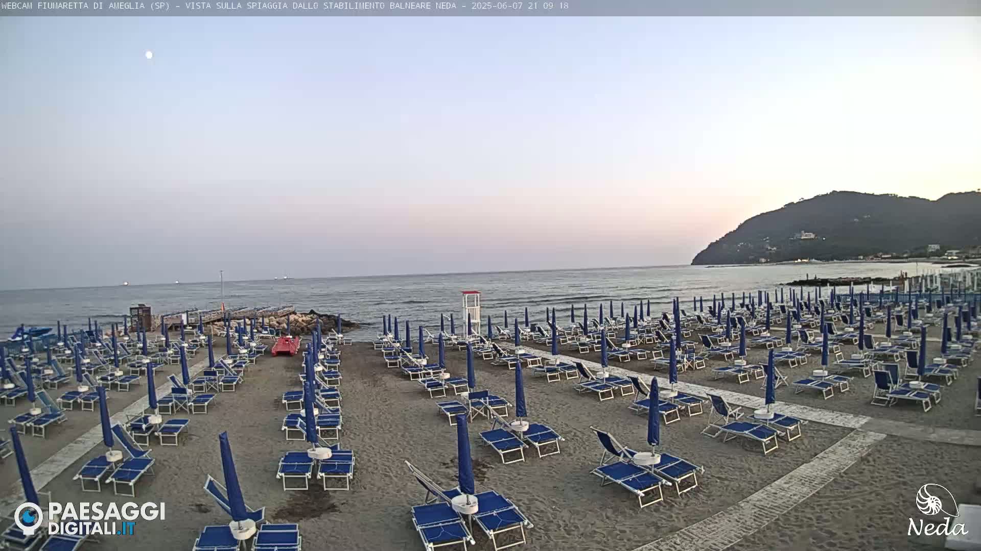 A beach with numerous rows of lounge chairs and umbrellas under a twilight sky, overlooking a calm sea with a distant hill.