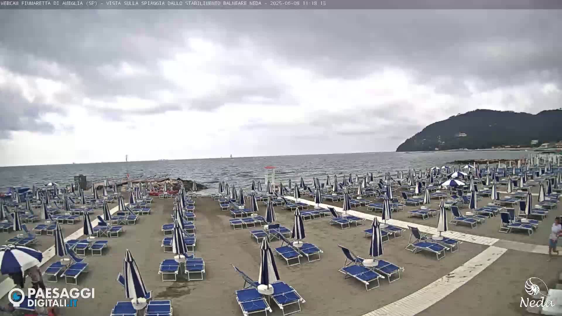 A mostly cloudy day at a beach with many rows of closed beach umbrellas and lounge chairs on a sandy shore, and a distant hill visible across the water.
