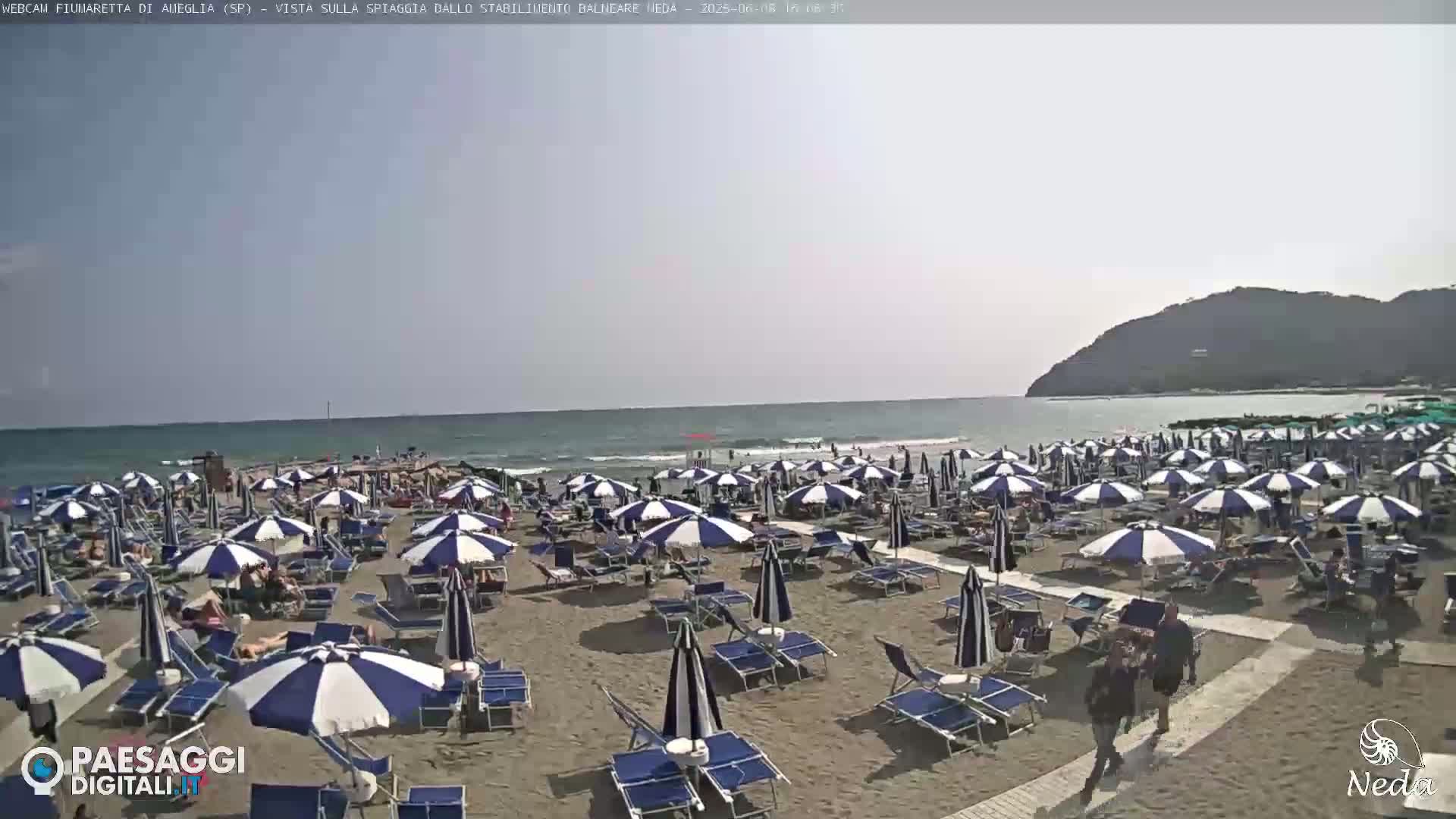 A sandy beach with numerous striped beach umbrellas and lounge chairs is visible, under a mostly clear sky, with a calm sea in the background.