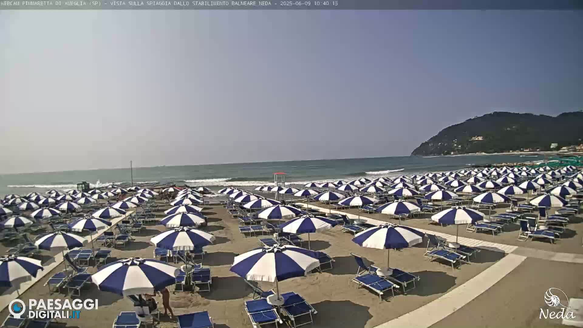 A sandy beach with many blue and white striped umbrellas and lounge chairs is situated next to a calm ocean under a clear sky.