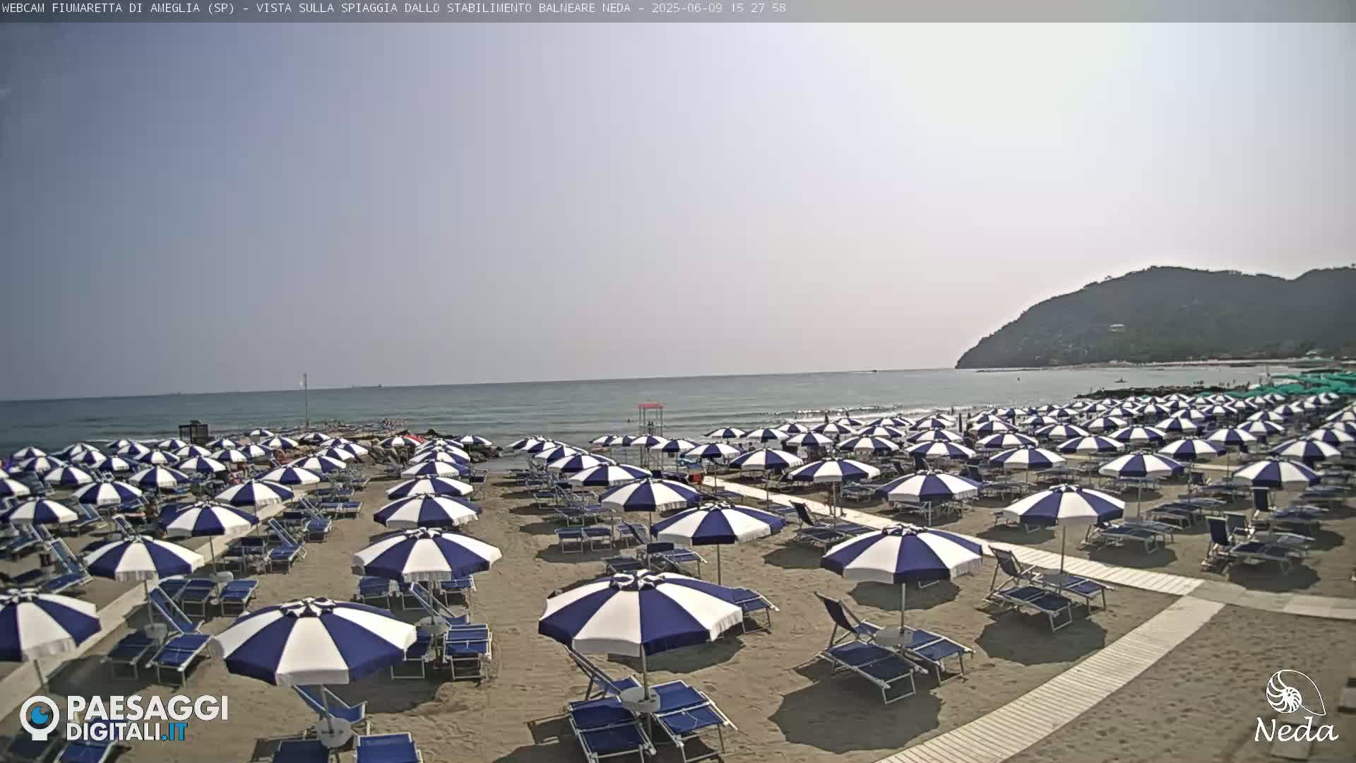 A sandy beach with numerous blue and white striped umbrellas and lounge chairs is situated next to a calm ocean under a hazy sky, and a hill is visible in the distance.