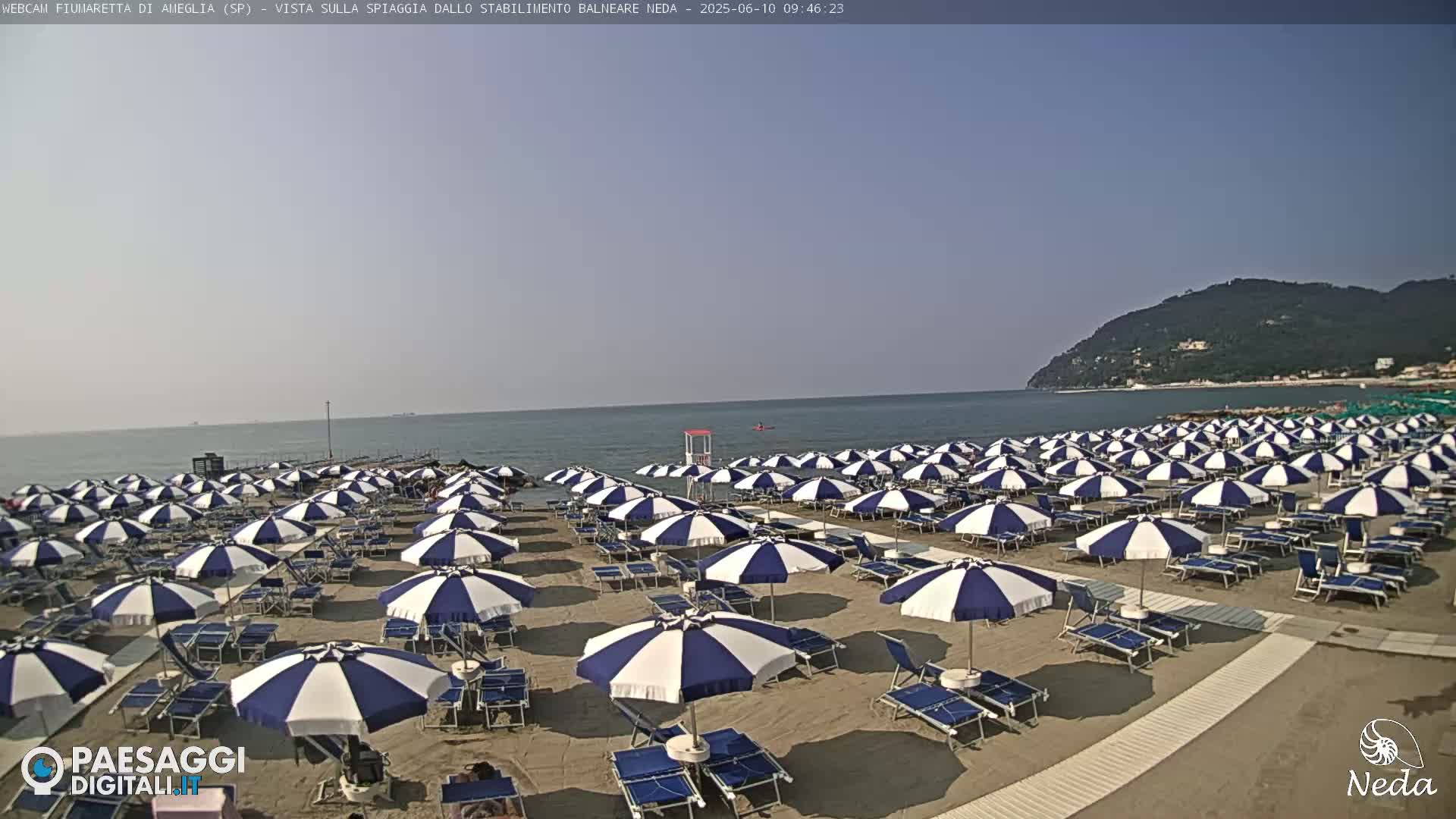 A sandy beach with numerous blue and white striped umbrellas and lounge chairs is situated by a calm ocean under a clear sky, with a distant hillside visible in the background.