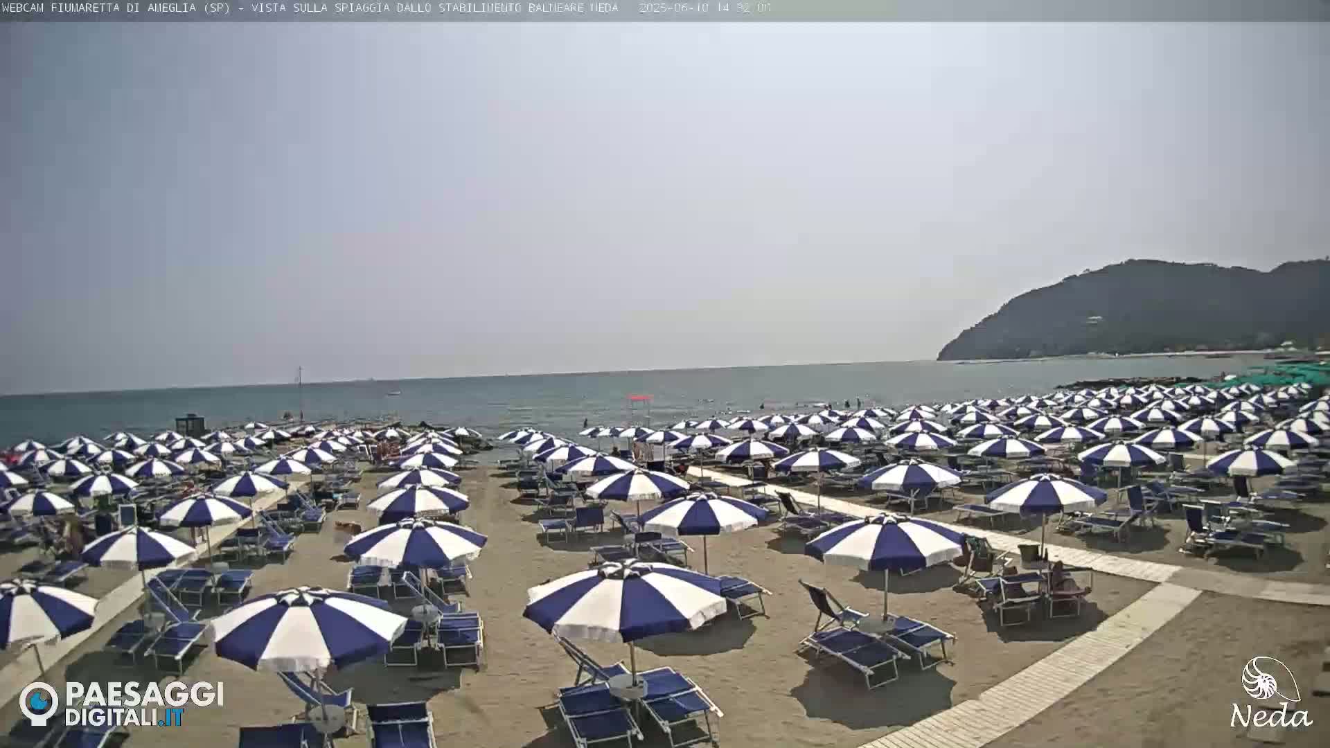 A sandy beach with many blue and white striped umbrellas and lounge chairs is visible under a sunny sky, next to a calm sea with a distant hill.