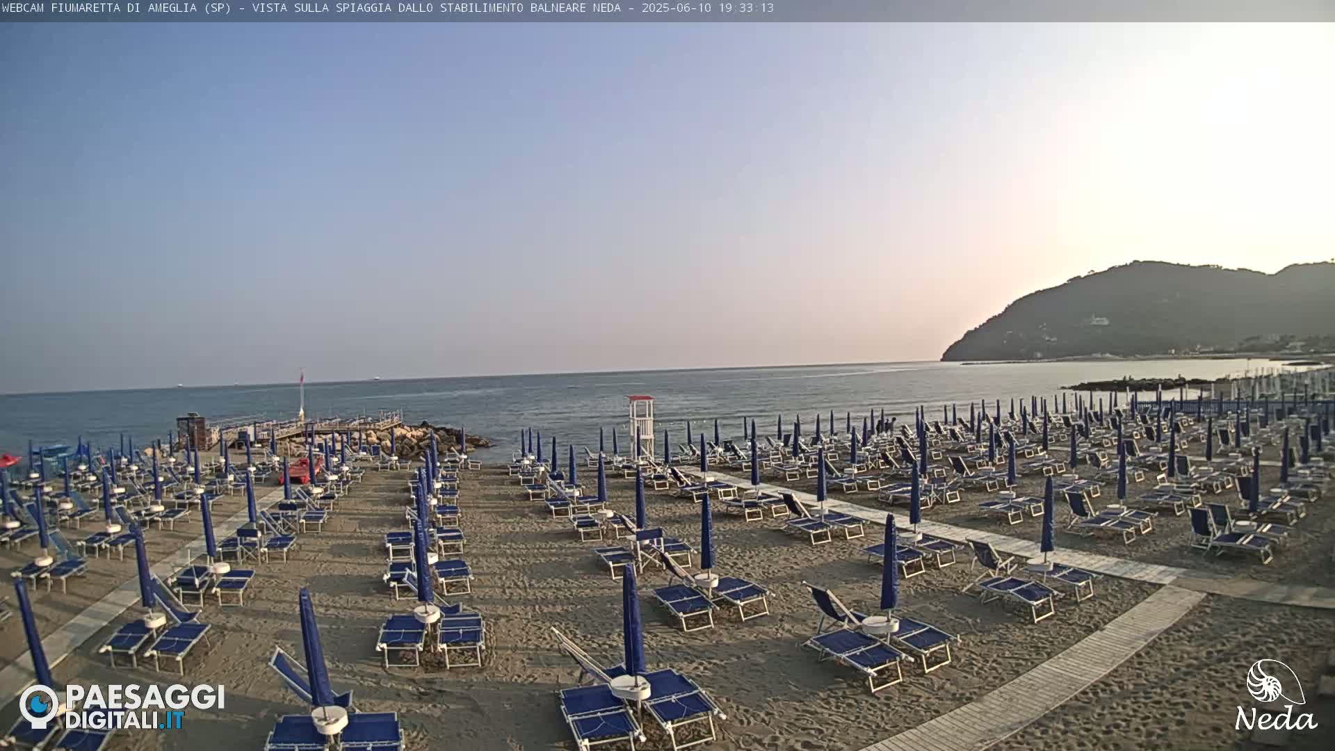 A mostly empty sandy beach with rows of blue beach umbrellas and lounge chairs is situated next to a calm ocean under a clear sky.