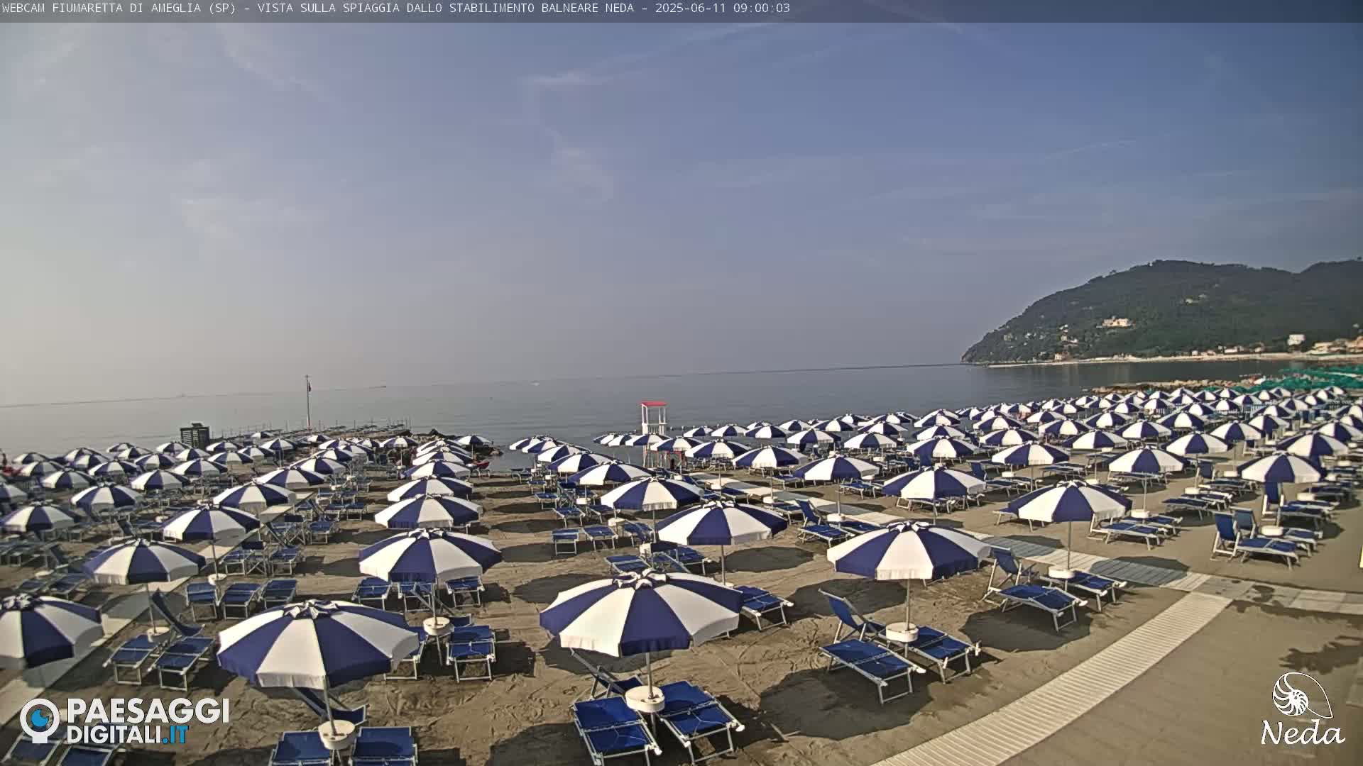 A sandy beach with numerous blue and white striped umbrellas under a clear sky, overlooking a calm sea and a distant hillside.