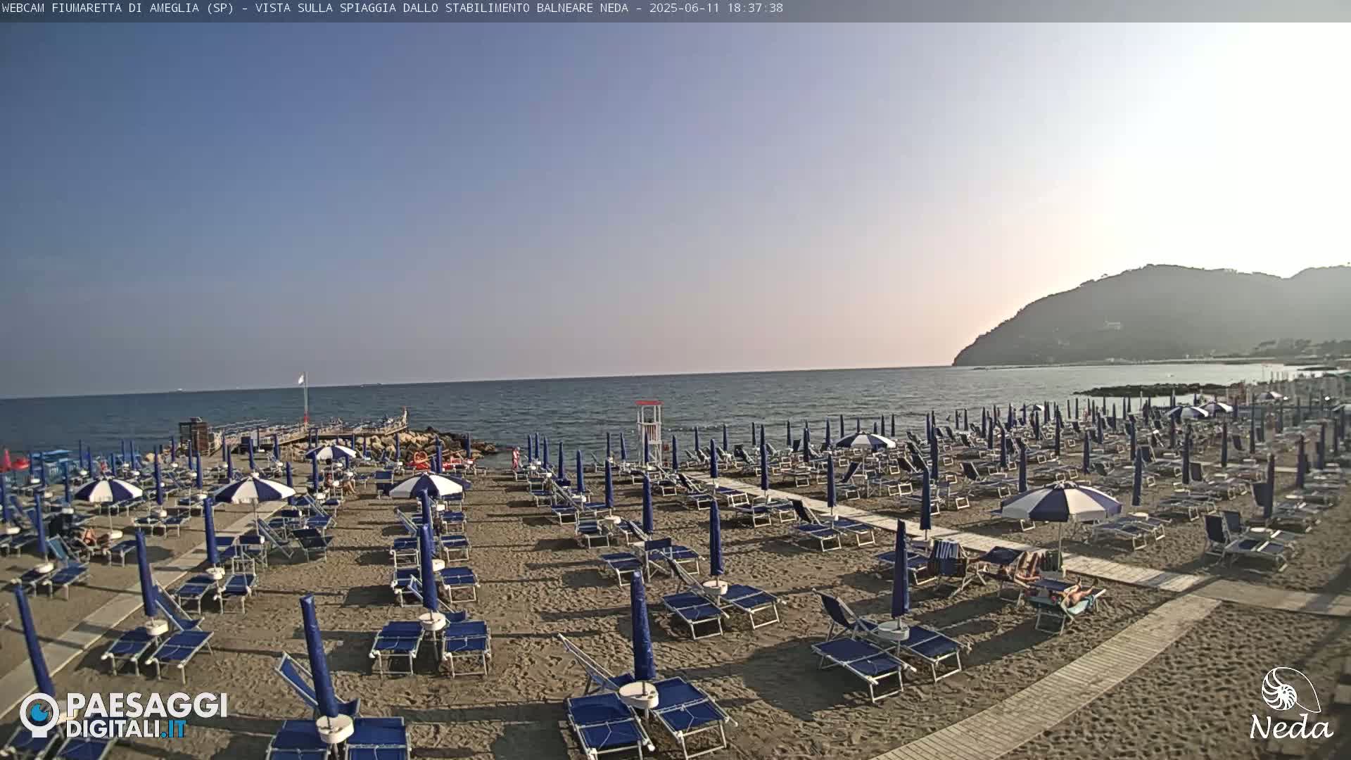 A sandy beach with numerous blue and white umbrellas and lounge chairs is situated on a calm sea under a clear, sunny sky, with a hill visible in the background.