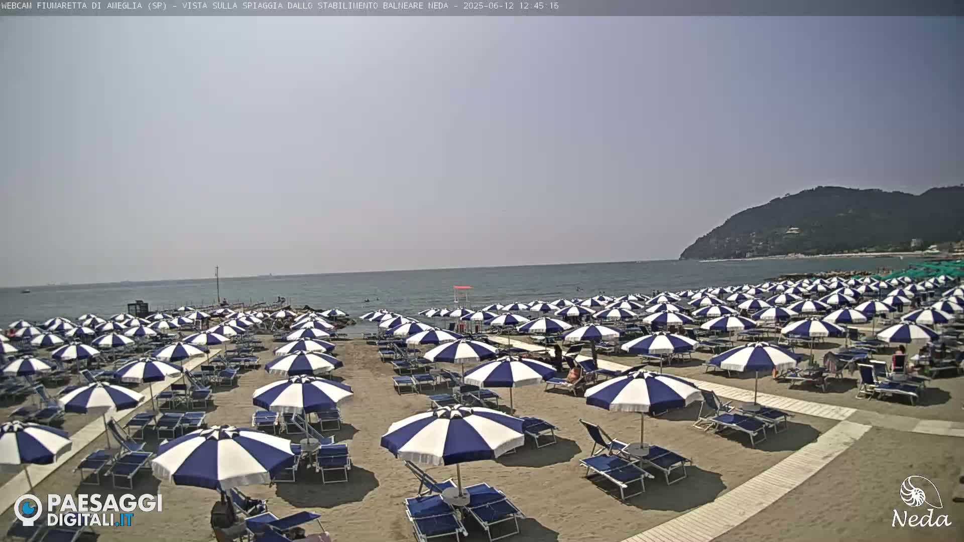 A sandy beach with numerous blue and white striped umbrellas under a clear, sunny sky, next to a calm sea and a hill in the distance.