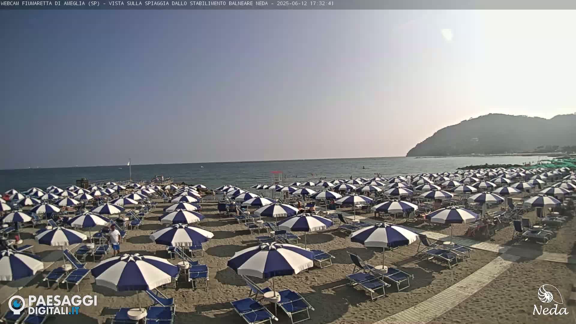 A sandy beach with numerous blue and white striped umbrellas and lounge chairs is situated next to a calm sea under a clear, sunny sky, with a distant hill visible in the background.
