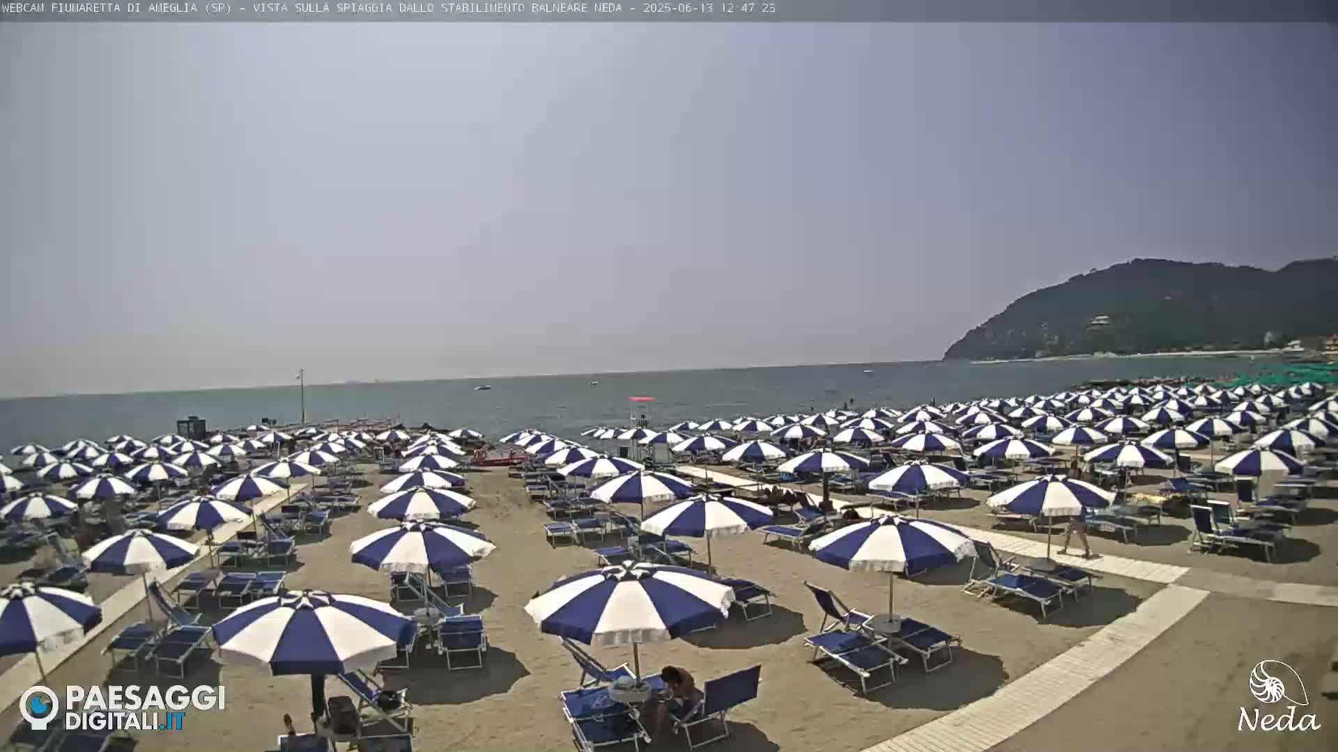 A sandy beach with numerous blue and white striped umbrellas and lounge chairs stretches towards a calm ocean under a clear sky.