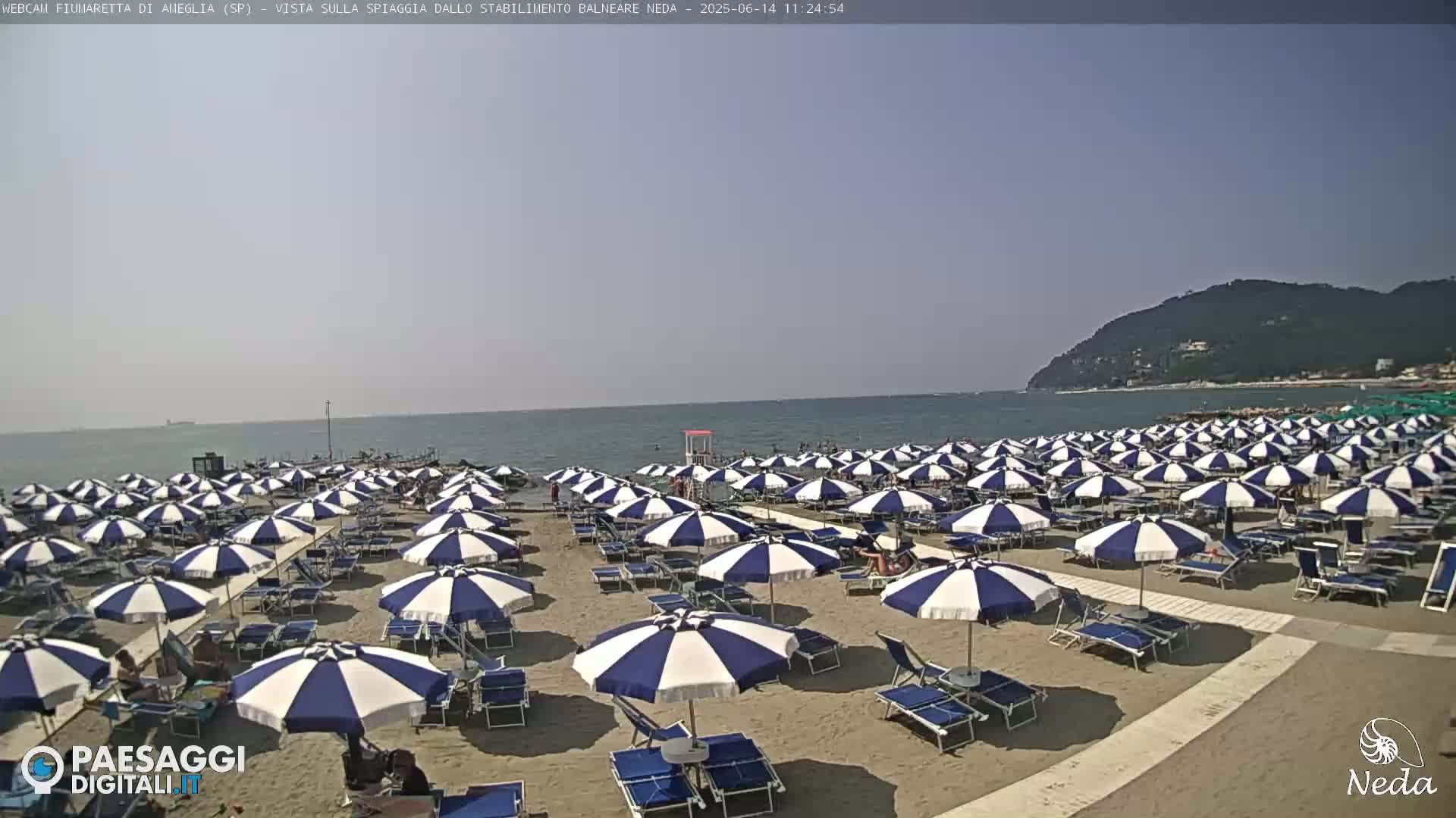A sandy beach filled with numerous blue and white striped umbrellas and lounge chairs stretches toward a calm ocean under a clear sky, with a distant hillside visible.