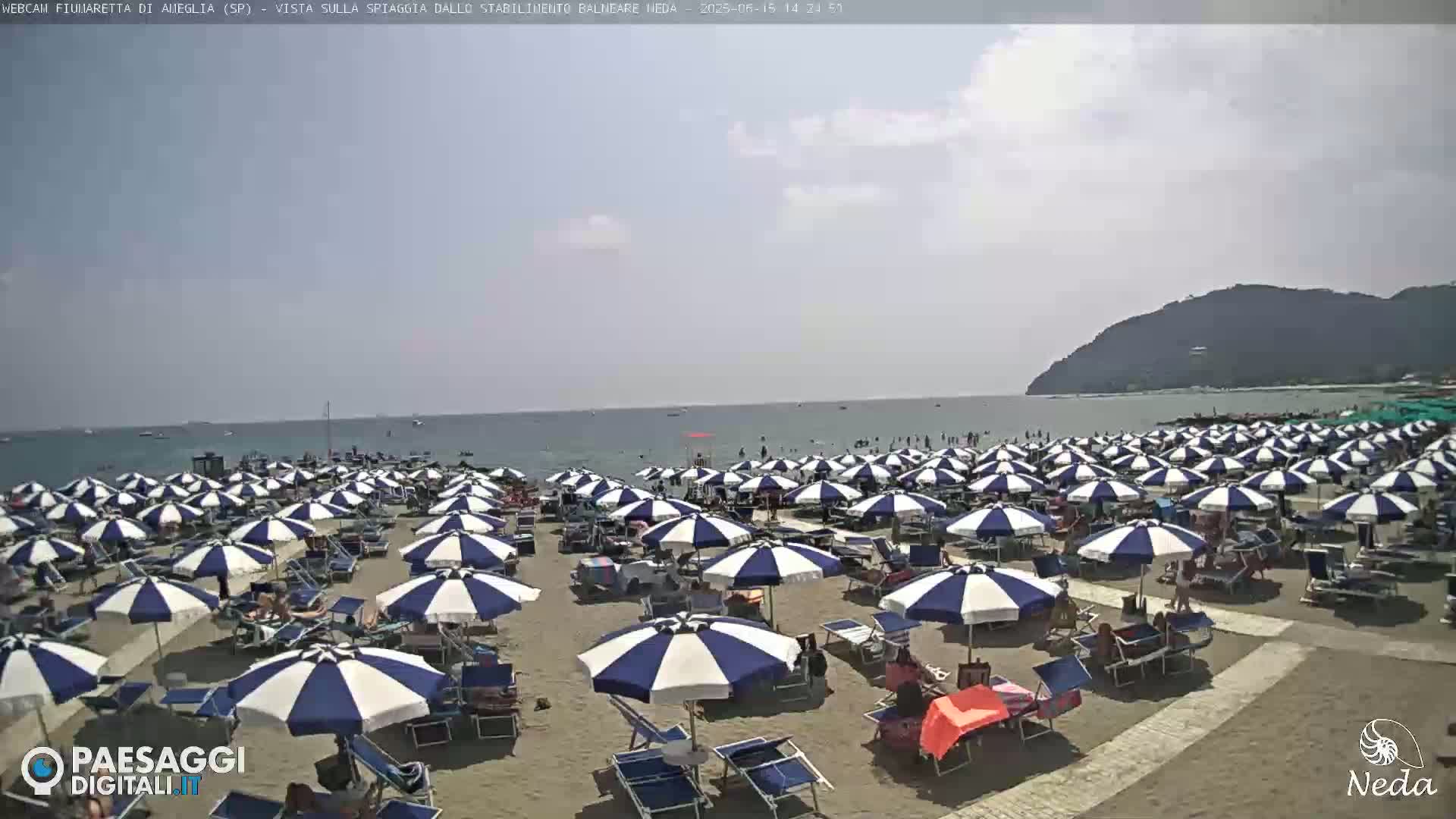A sandy beach is crowded with numerous blue and white striped beach umbrellas under a partly sunny sky, with calm ocean water visible in the background.