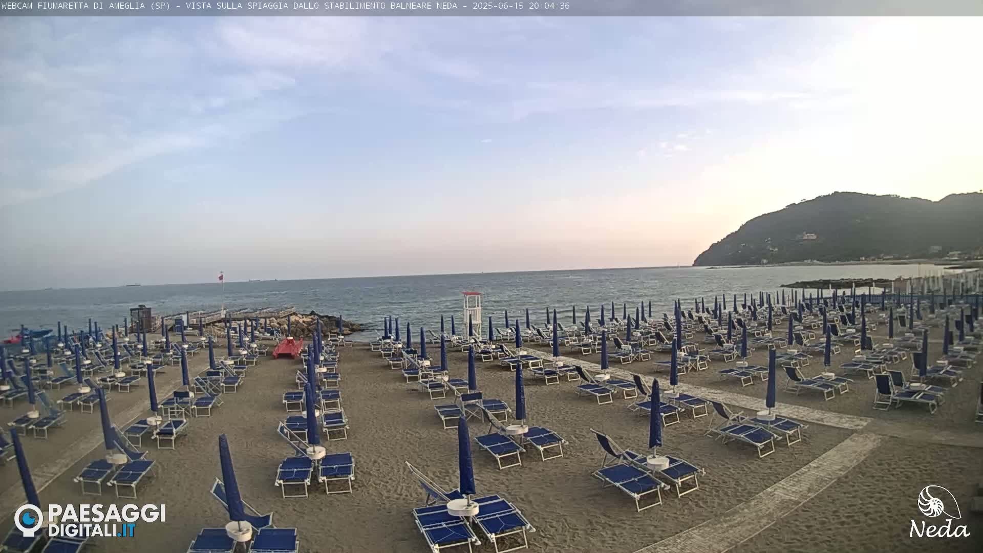 A sandy beach with numerous blue umbrellas and lounge chairs sits under a partly cloudy twilight sky, overlooking a calm sea and a distant hill.