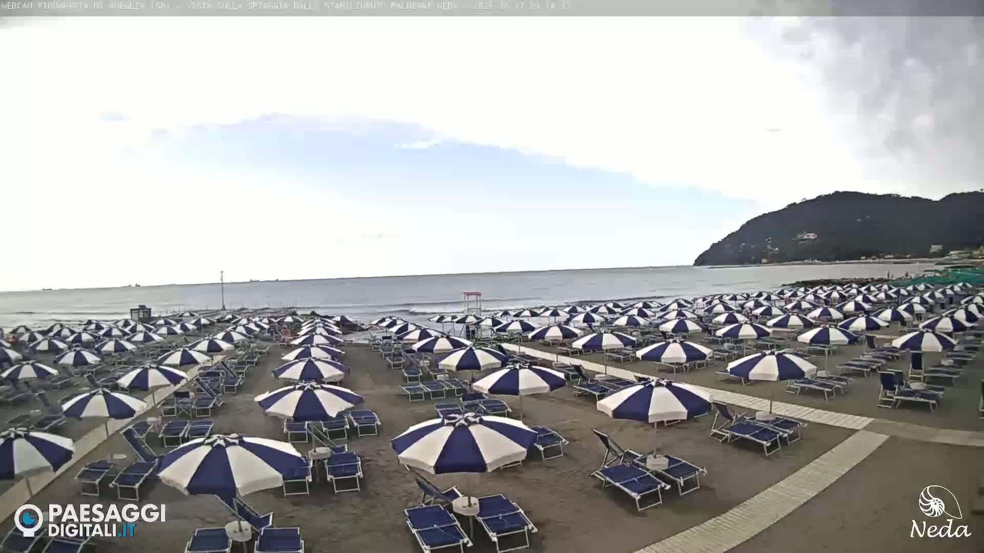 A sandy beach with numerous blue and white striped umbrellas and lounge chairs is visible, overlooking a calm ocean under an overcast sky.