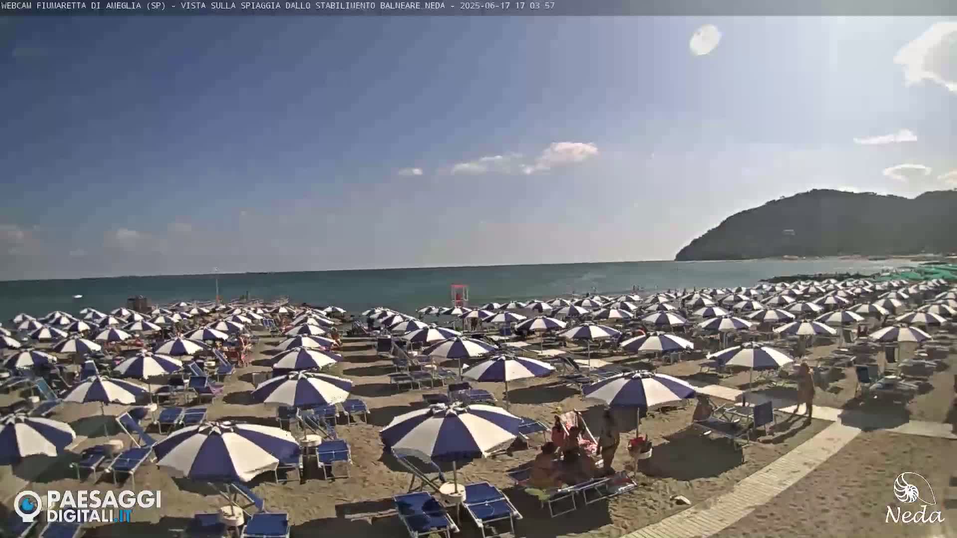 A sandy beach with numerous blue and white striped umbrellas and lounge chairs under a mostly sunny sky, next to a calm ocean with a hill in the background.