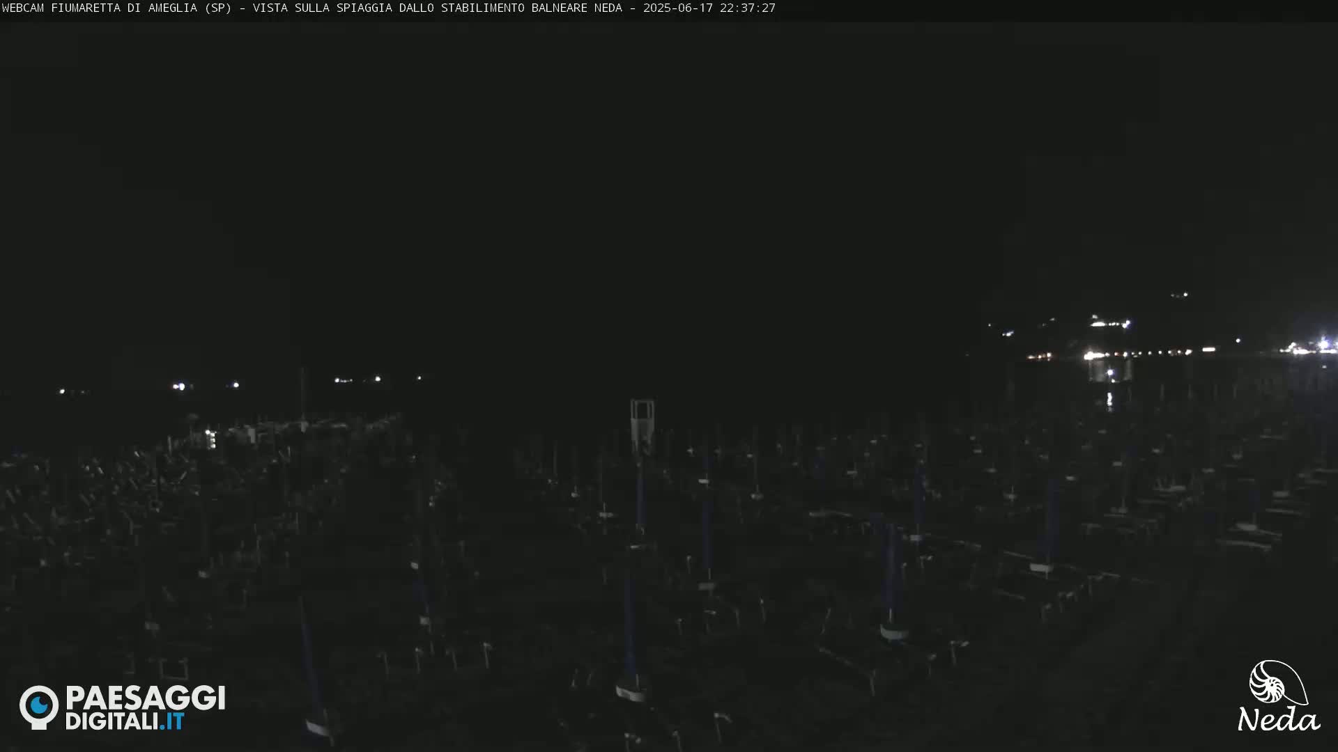 A dark beach at night is shown, with many closed beach umbrellas and a faintly visible shoreline in the distance.