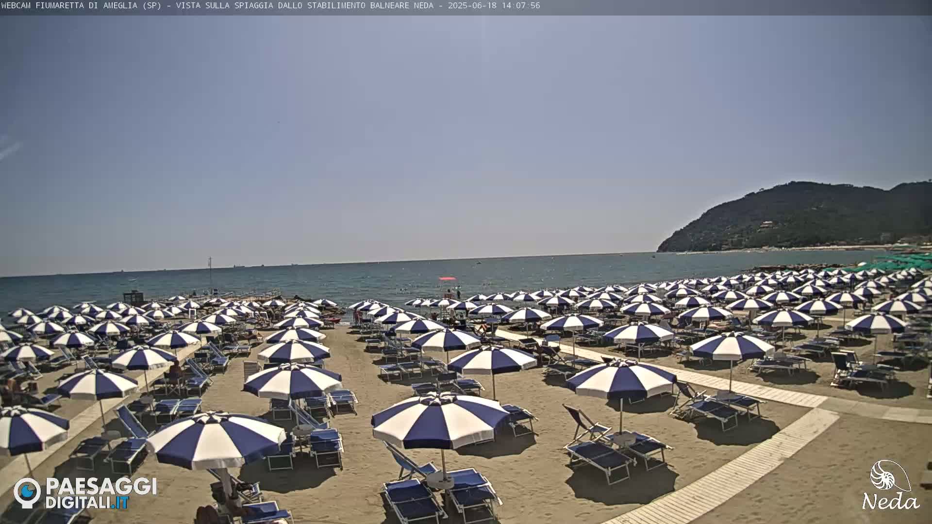 A sandy beach with numerous blue and white striped umbrellas under a clear, sunny sky, backed by a calm ocean and a distant hill.