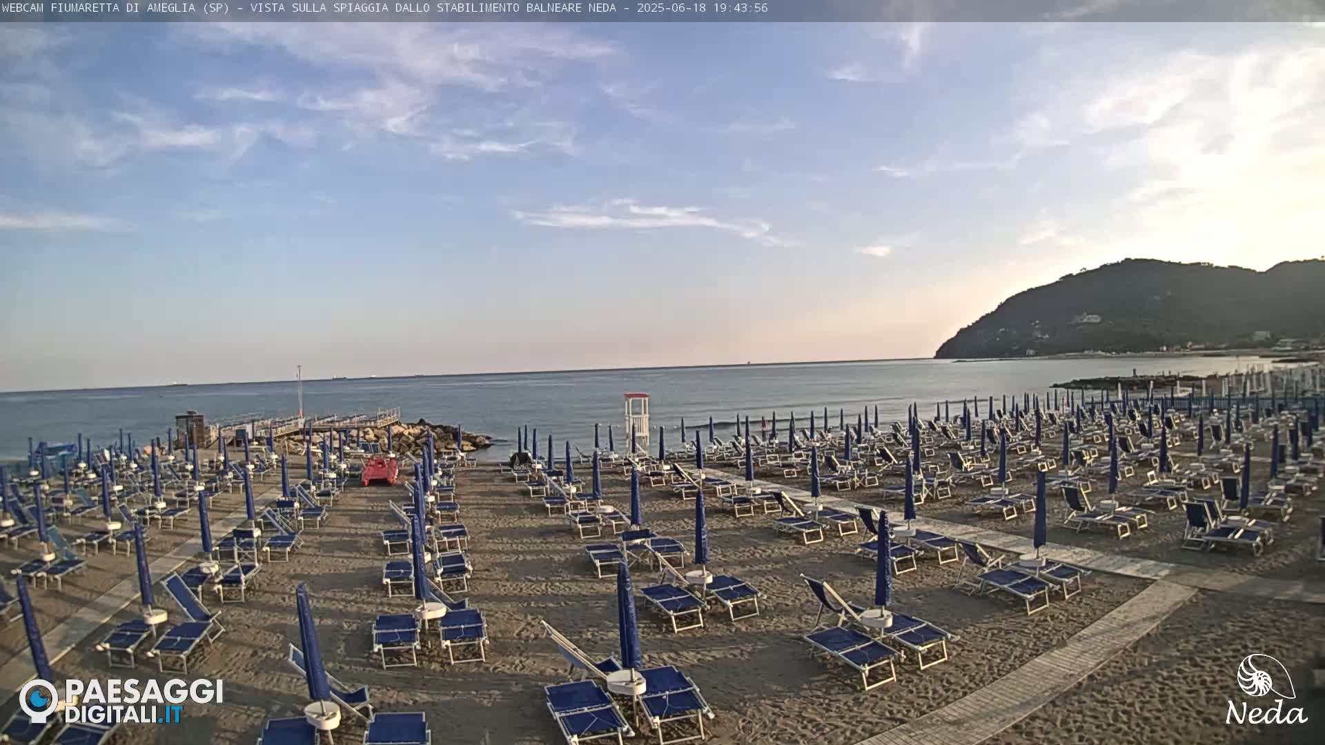 A sandy beach with numerous blue umbrellas and lounge chairs is situated next to a calm ocean under a partly cloudy sky.