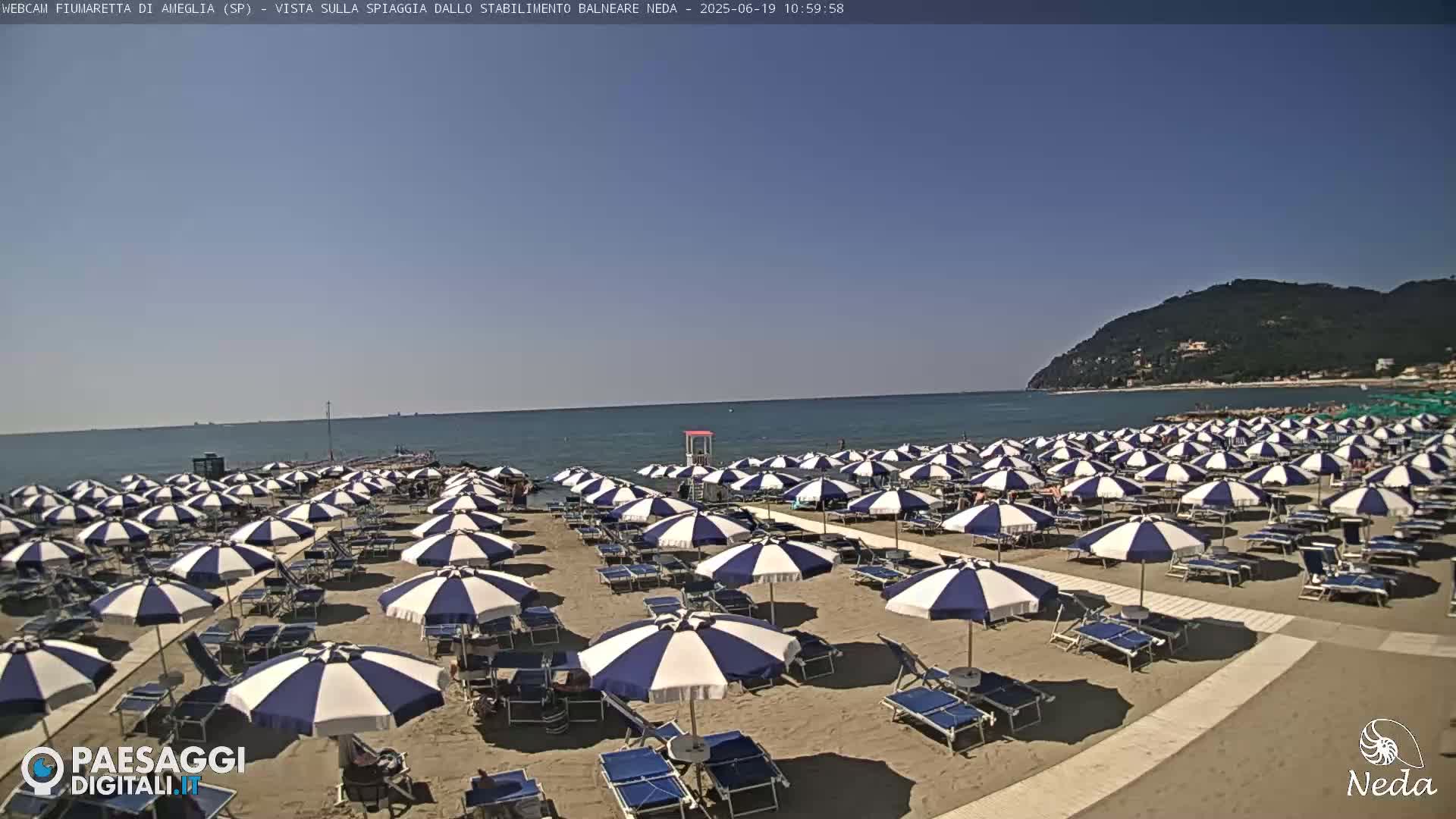 A sandy beach with numerous blue and white striped umbrellas and lounge chairs is visible on a sunny day, next to a calm ocean and a distant mountain range.