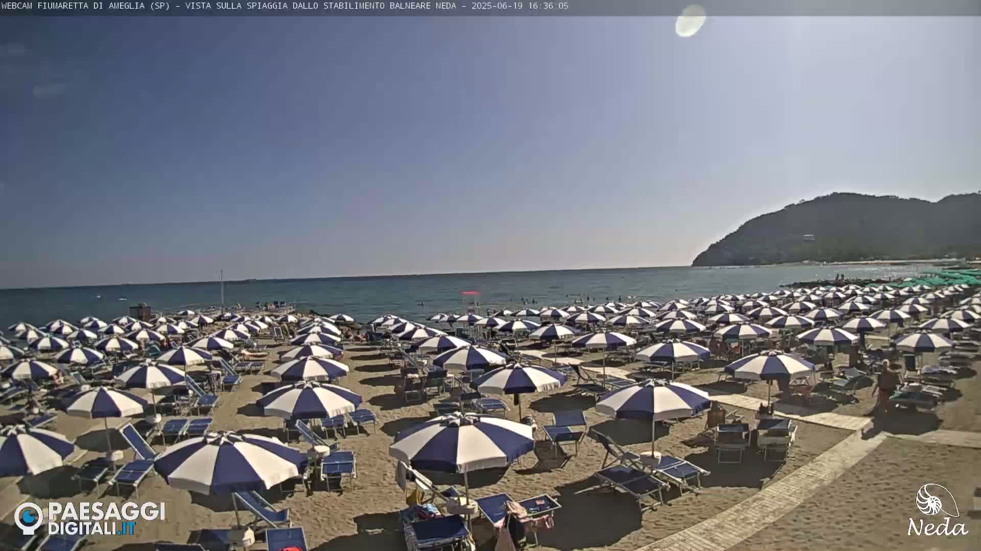 A sandy beach with numerous blue and white striped umbrellas and beach chairs is situated on a calm ocean under a clear blue sky.