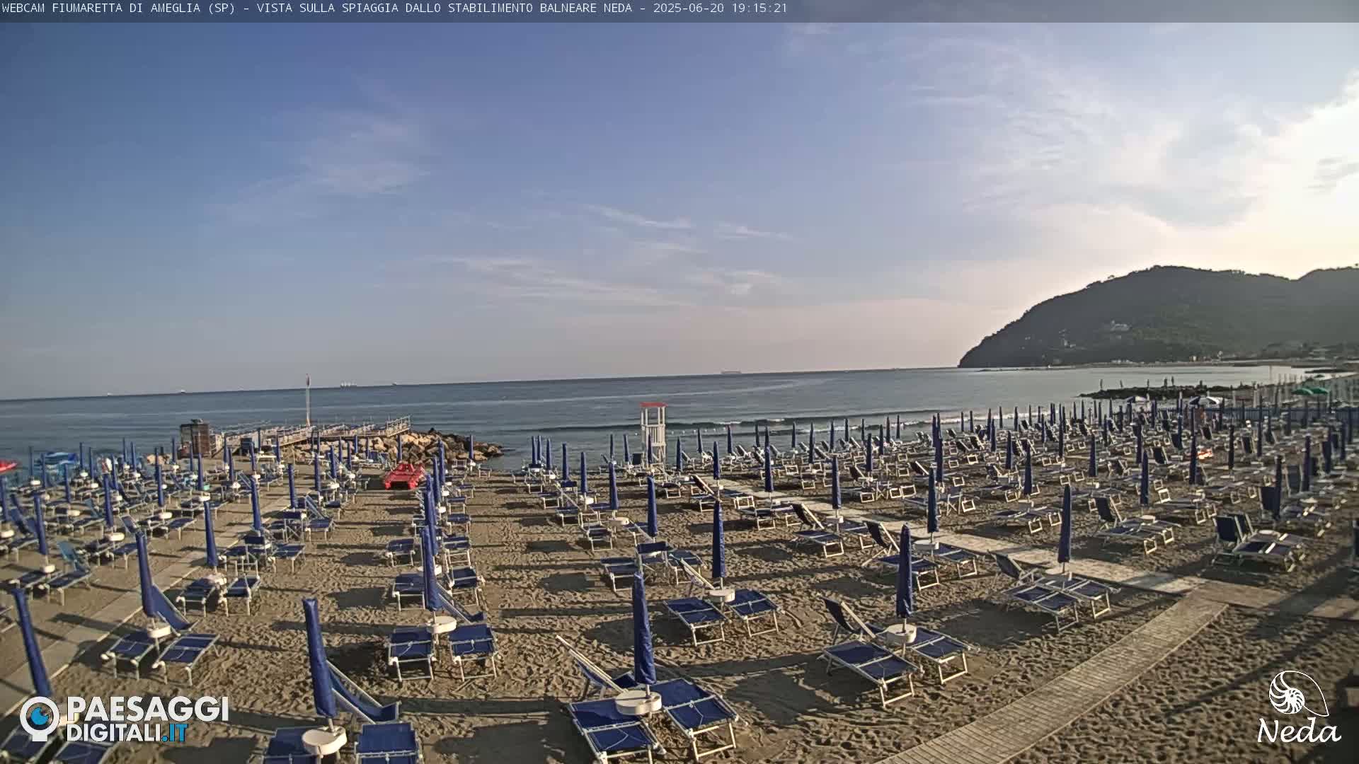 A sandy beach with rows of blue umbrellas and lounge chairs is situated next to a calm ocean under a mostly clear sky, with a distant hill visible in the background.