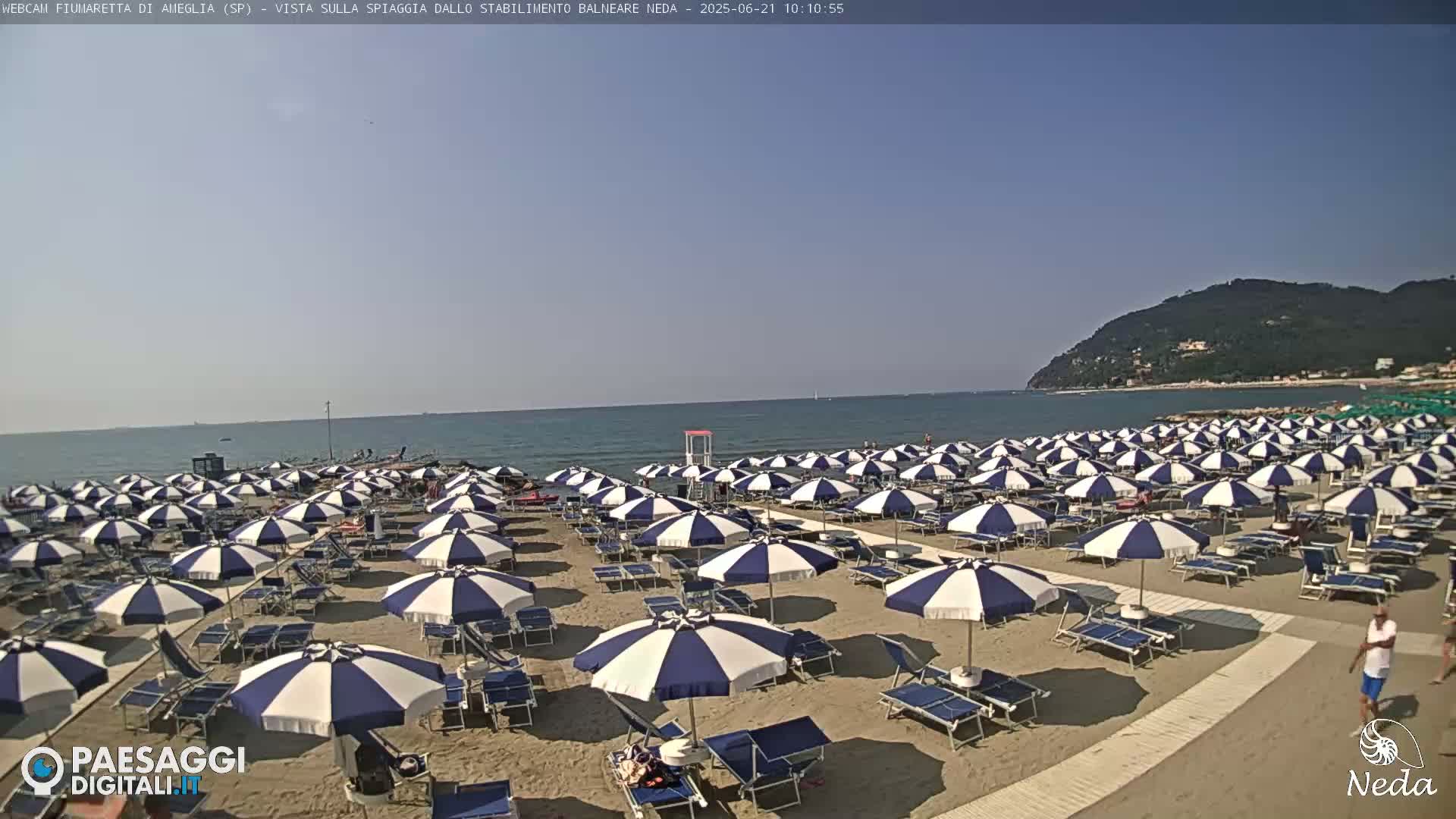 A sandy beach with numerous blue-and-white striped umbrellas and lounge chairs under a clear, sunny sky, overlooking a calm ocean and a distant hillside.