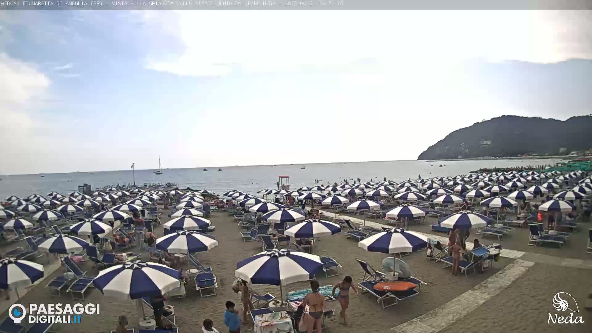 A sandy beach is crowded with numerous blue and white striped beach umbrellas under a partly cloudy sky, next to a calm sea.