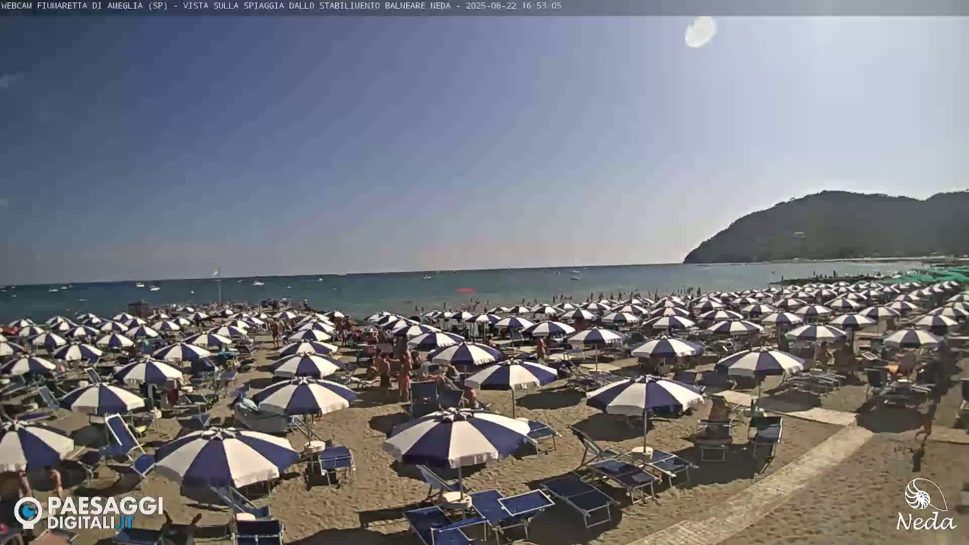 A sunny beach scene shows many blue-and-white striped umbrellas and beach chairs on a sandy beach next to a calm ocean with a hill in the background.