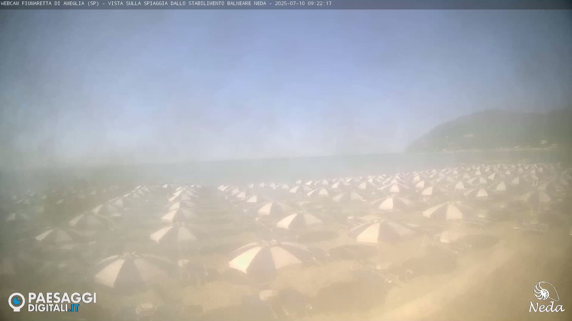 A hazy beach scene shows numerous beach umbrellas on a sandy shore near a calm sea and a distant shoreline.